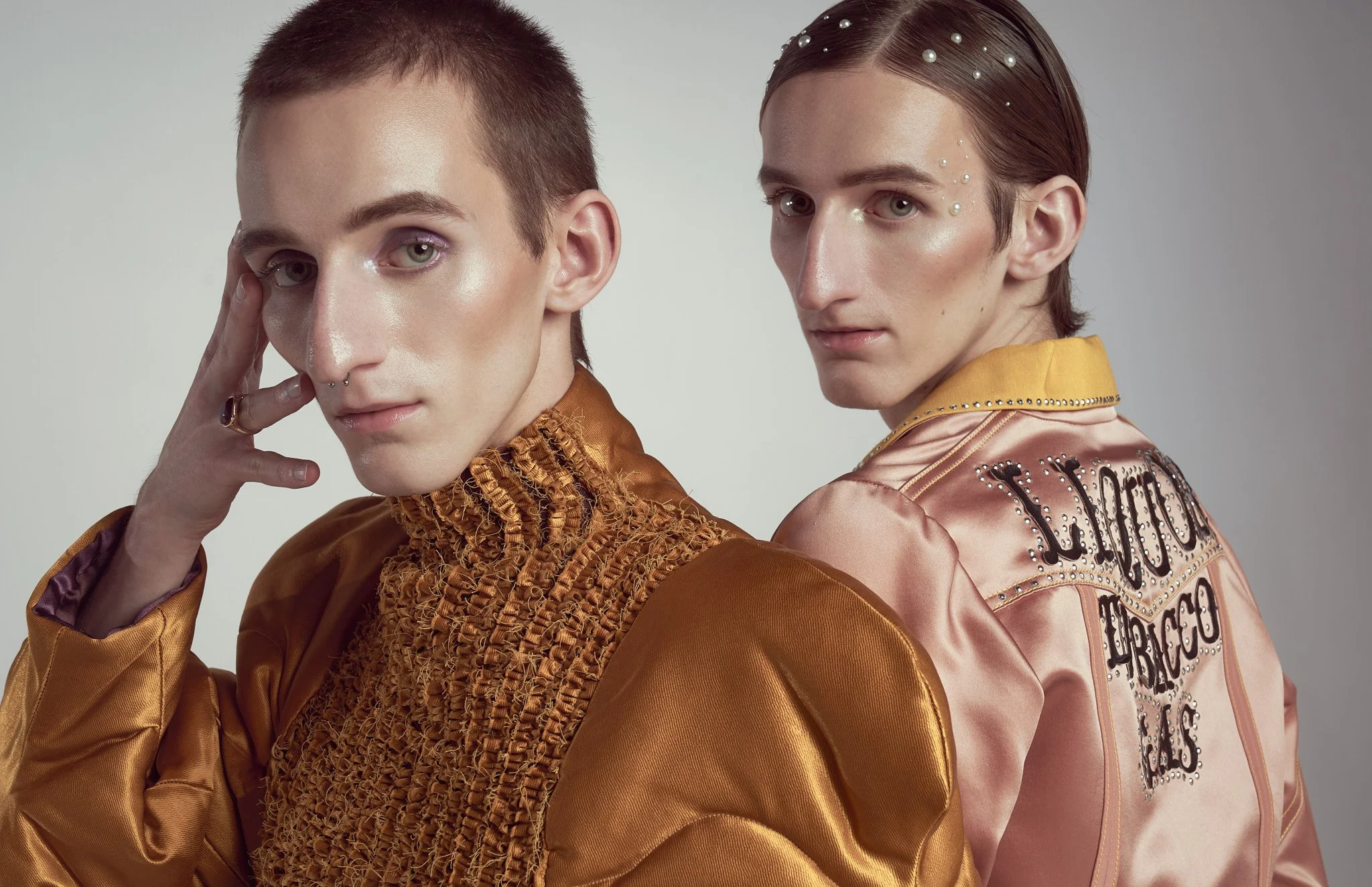 Half-length editorial photo of twin male models, one wearing a golden smocked high-neck top and the other wearing a satin jacket with pearl makeup detailing.⁠