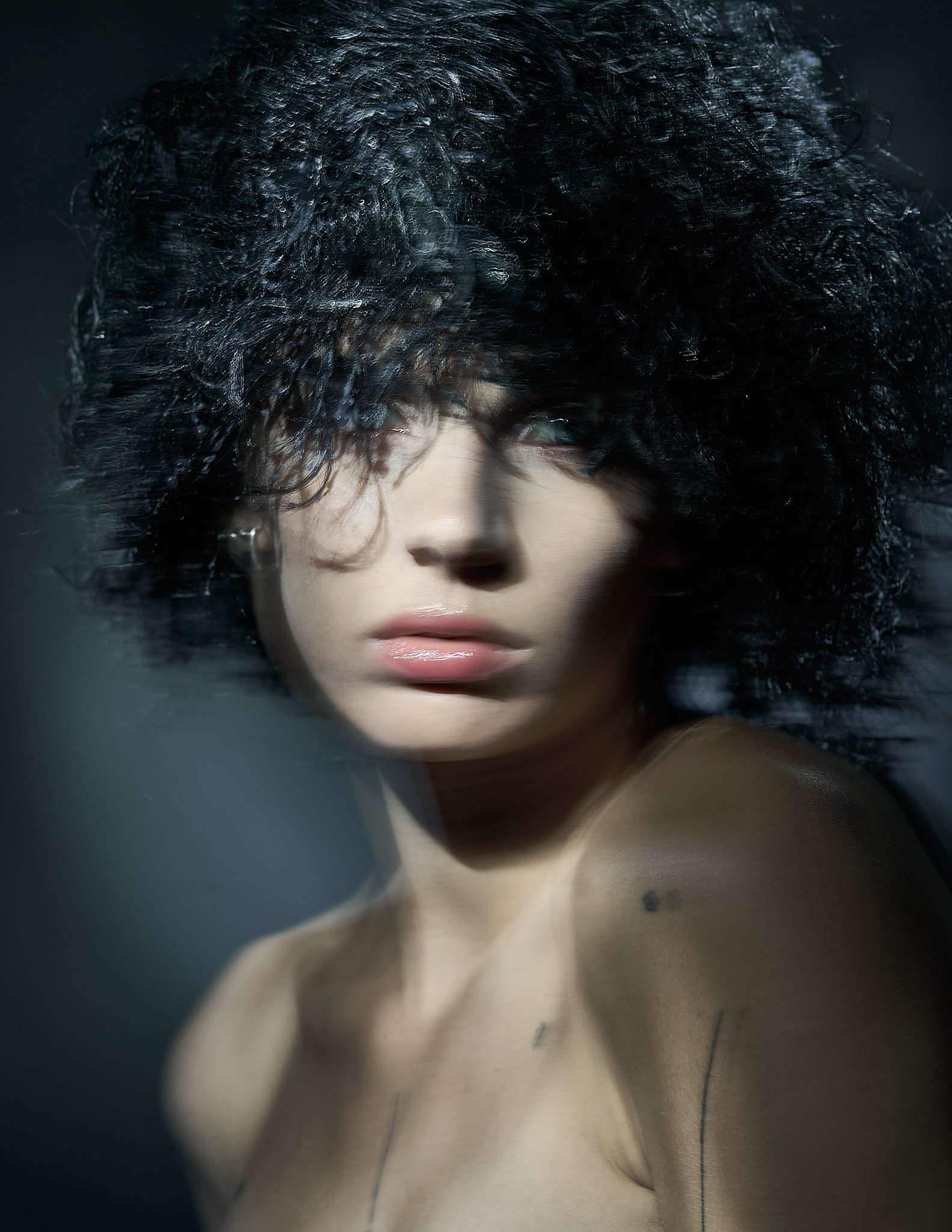 Avant-garde beauty portrait of a model with voluminous, textured black fringe wig covering the forehead, highlighted by dramatic studio lighting.⁠