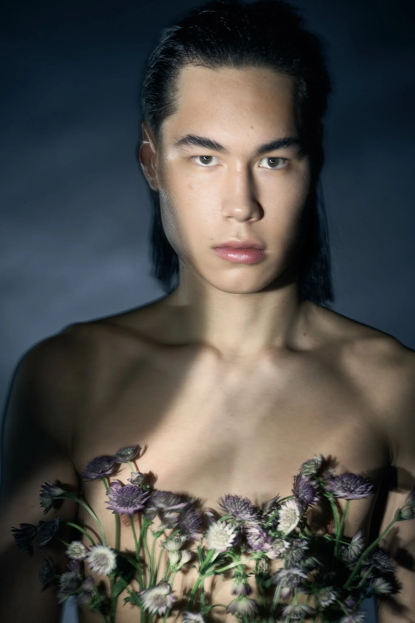 Editorial beauty portrait of a male model with long dark hair and sculptural lighting, photographed by Belgian fashion photographer Simon Rogghe in Ghent. The model poses shirtless with dried wildflowers arranged across the chest, creating a poetic, 