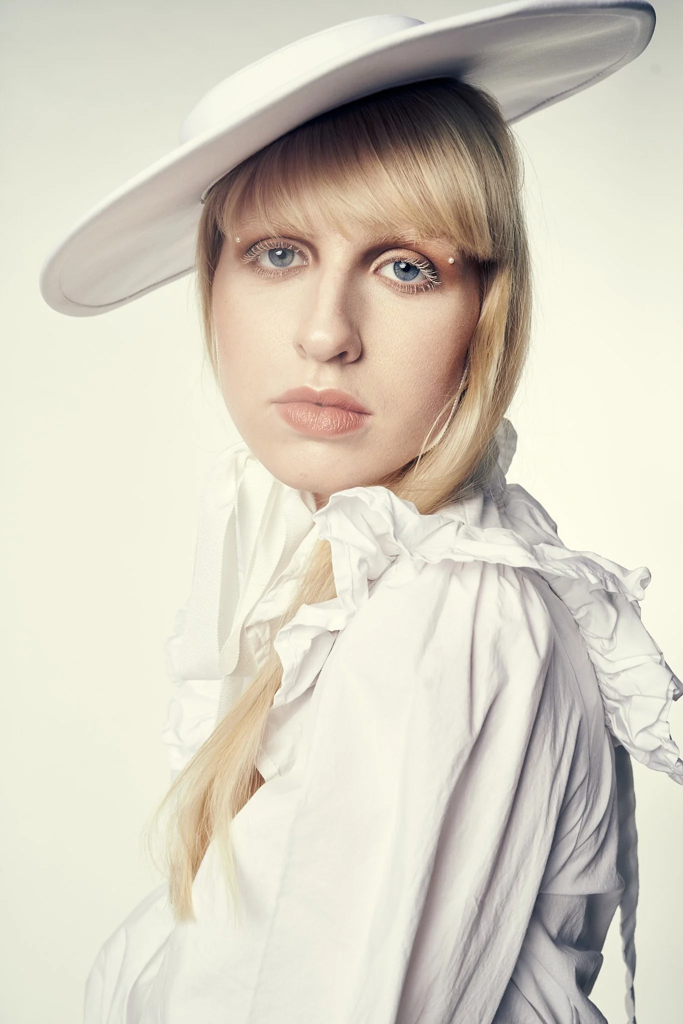 Close-up fashion portrait of a blonde model wearing a wide-brim white hat and a high-neck white ruffled blouse against a clean background.⁠