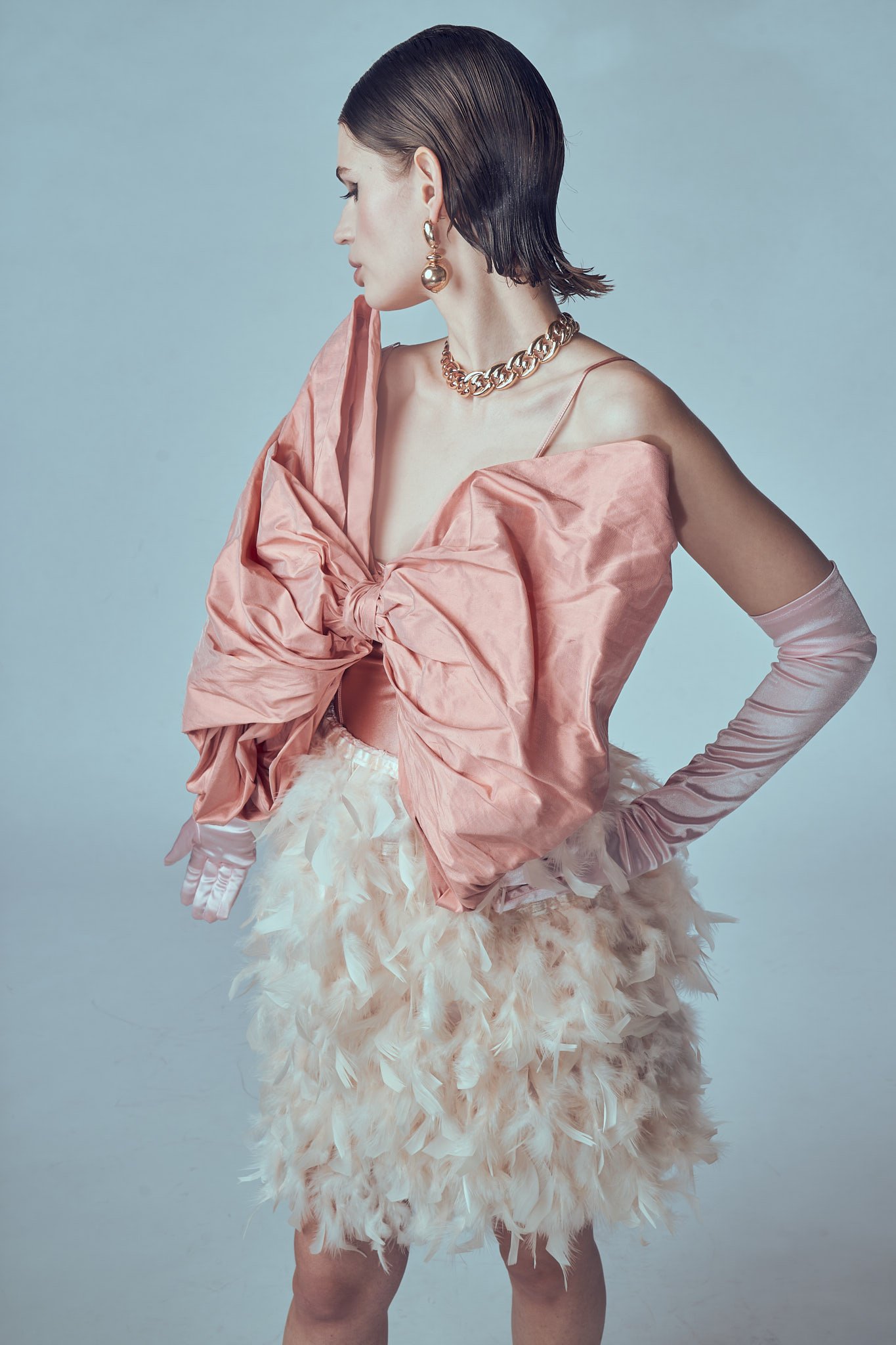 Studio fashion shot of a dark-haired model wearing an oversized pink bow blouse, long gloves, and a gold chain necklace.⁠