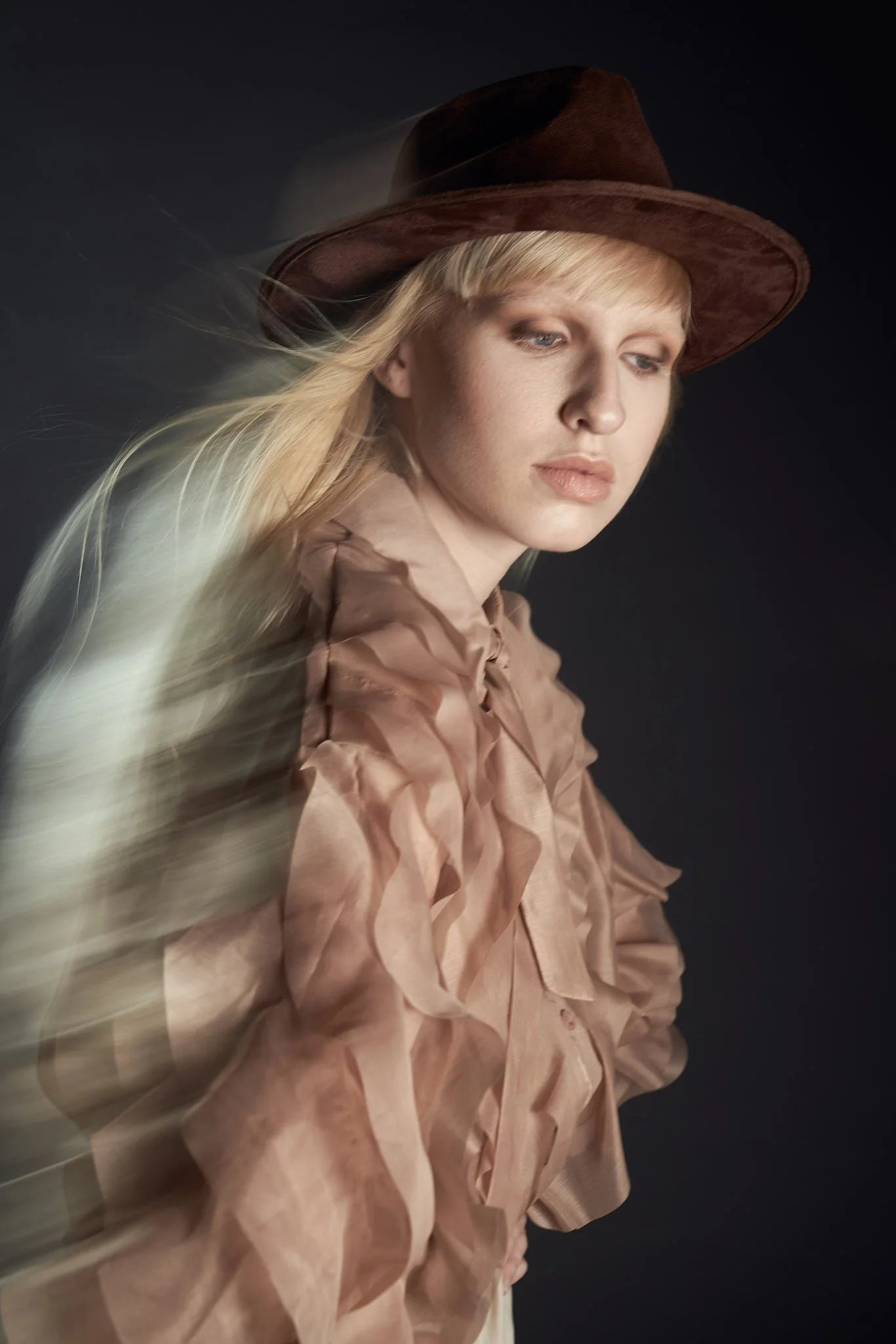Profile beauty portrait of a blonde model wearing a brown Western hat and a ruffled tan shirt with motion blur hair effect.⁠