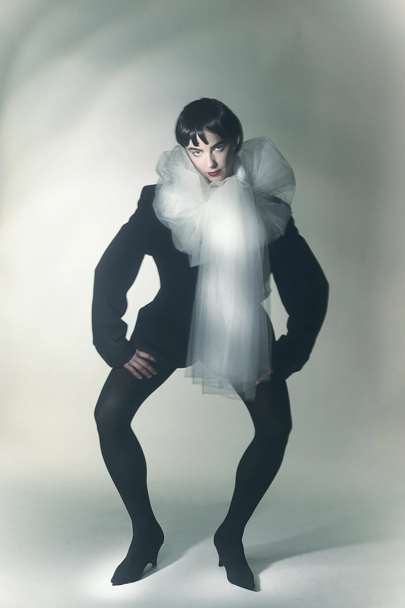 Full-length editorial photo of a model in a dark blazer, black tights, and an oversized white tulle neck ruffle posing with bent legs.⁠