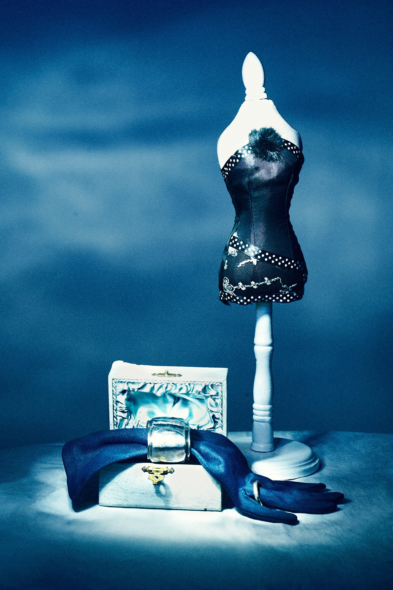 Commercial fine art still life showcasing luxury accessories, sculpted light techniques, and surreal composition with a miniature mannequin, silk glove, and silver jewelry on a moody blue backdrop.