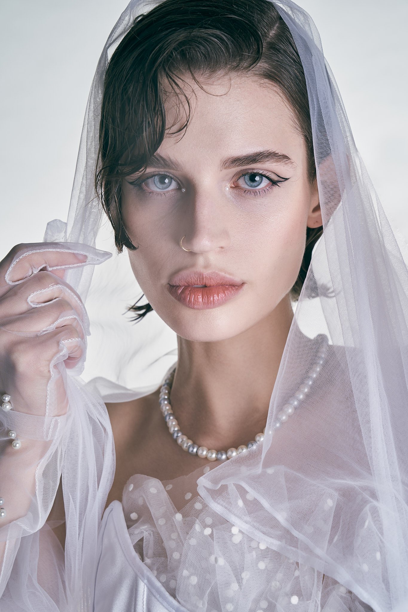 Editorial portrait of a model framed by sheer white netting, wearing a pearl necklace and a strapless embellished top.