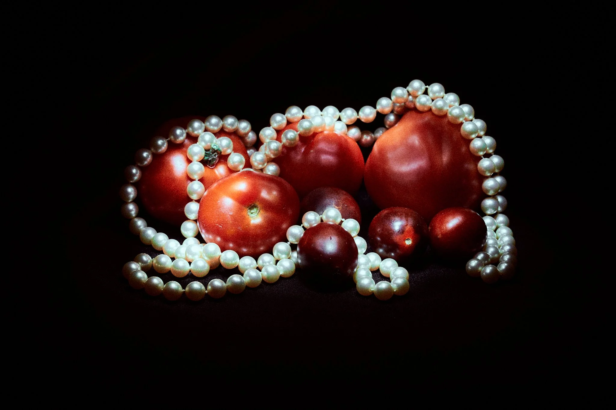 Still life photograph of dark red tomatoes intertwined with strands of white pearls set against a black background.