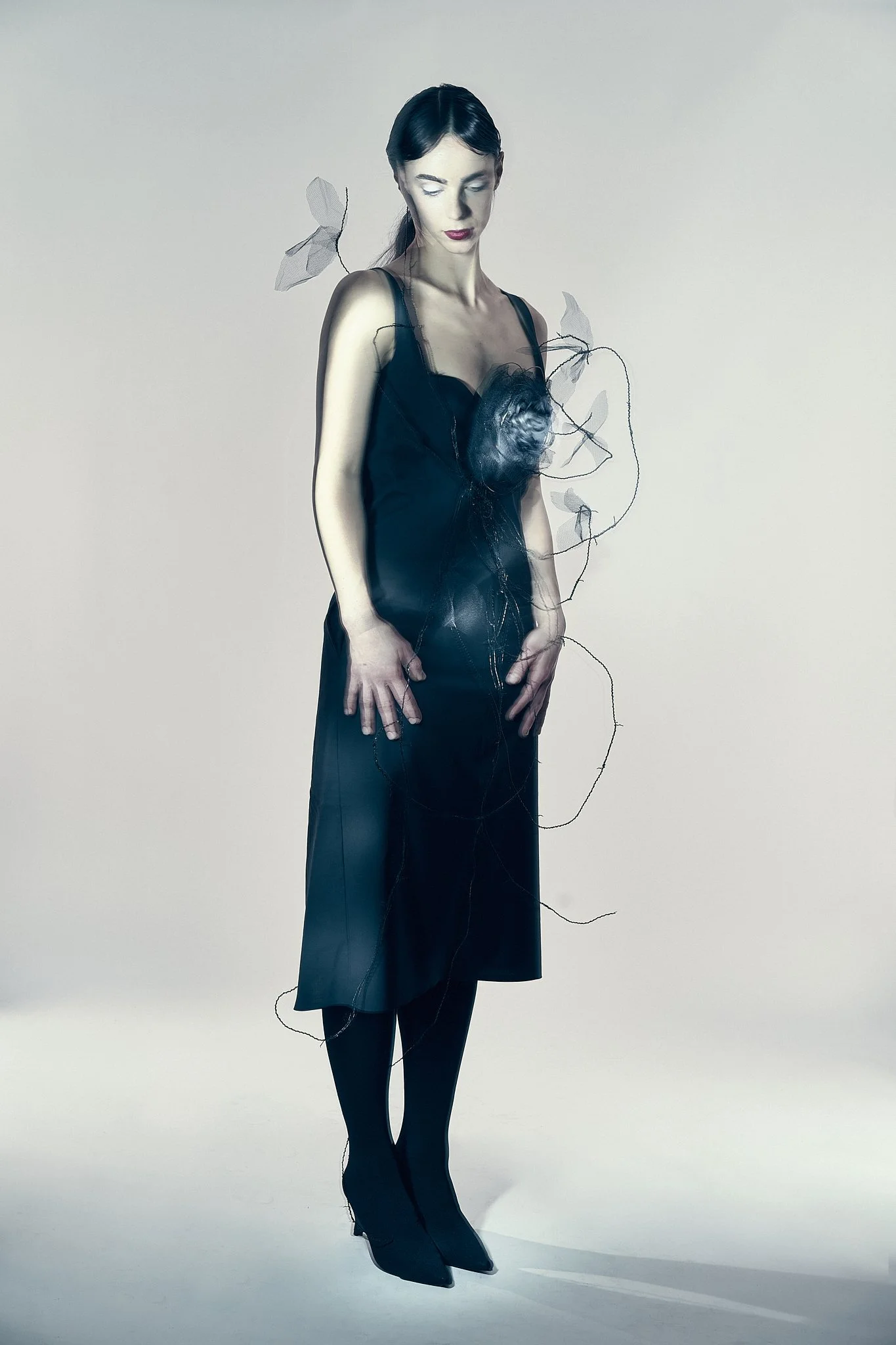 High-fashion studio portrait featuring a female model in a sleek black dress with avant-garde sculptural embellishments, captured with dramatic lighting for an editorial fashion photography campaign.