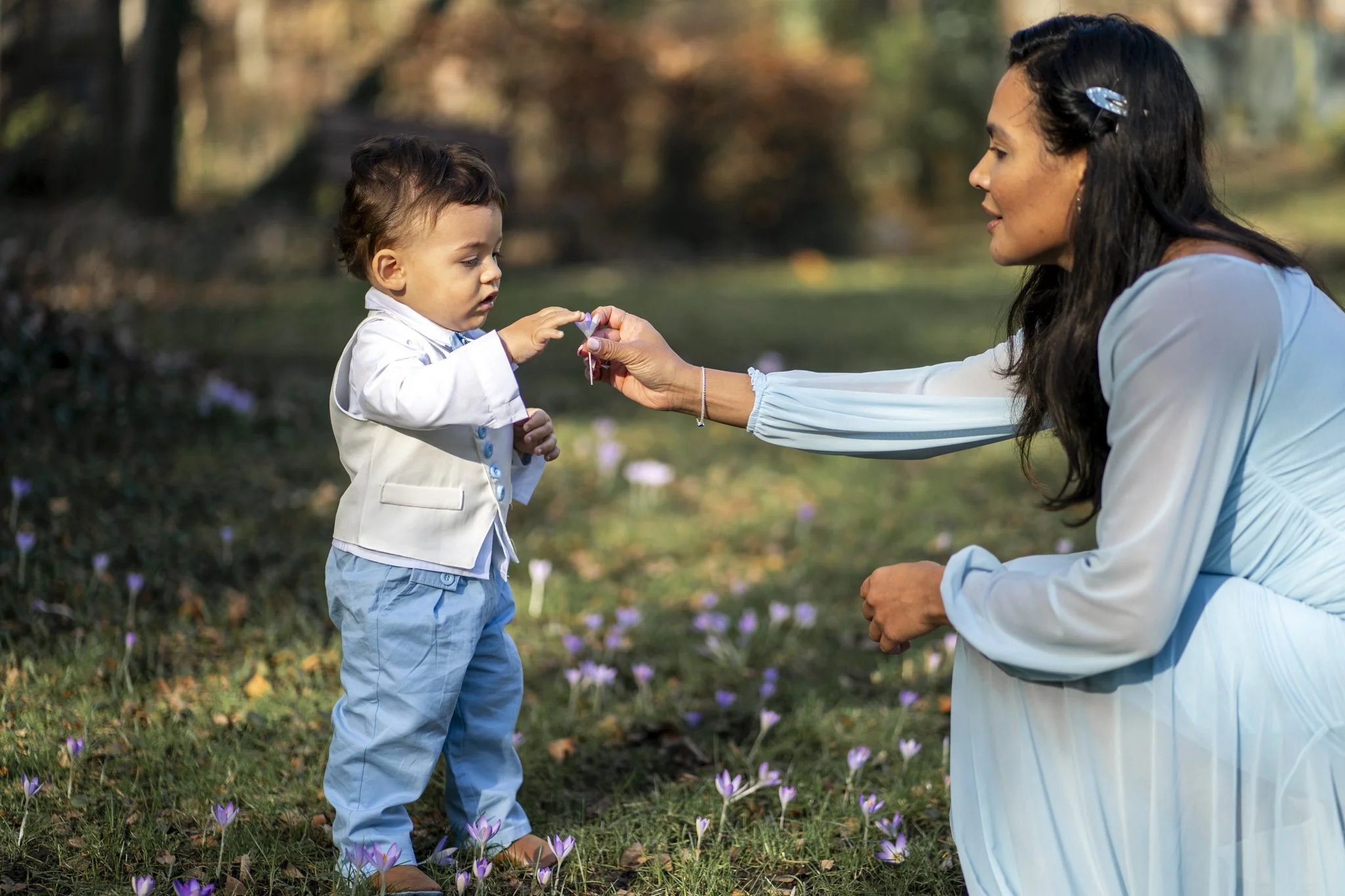 Moeder geeft bloemetje aan peuter tijdens buiten shoot – reportagefotografie Gent, natuurlijke kinderfoto’s.