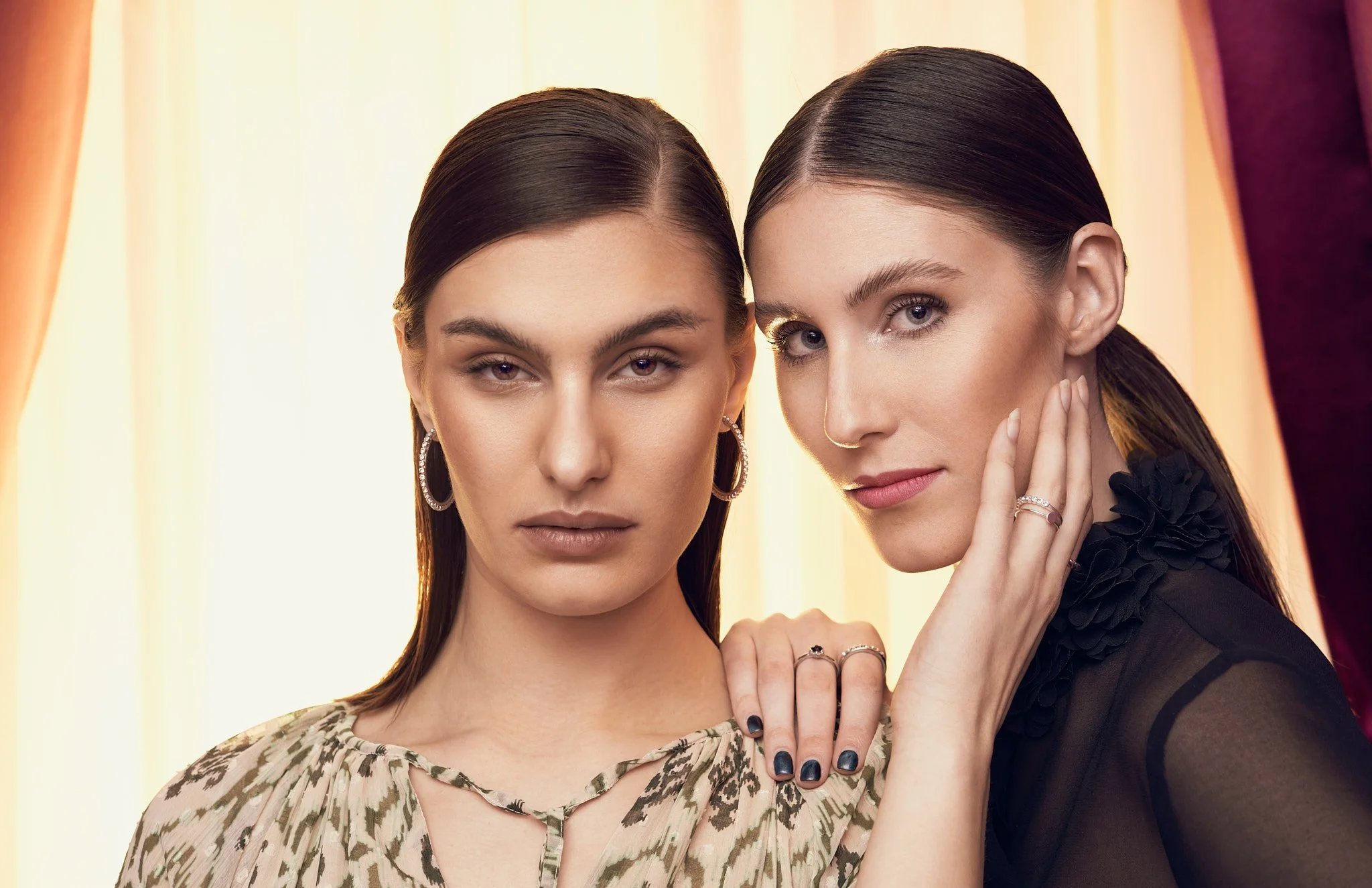 Beauty portrait of two dark-haired models posing close together against a soft lit background in high-neck vintage tops.⁠