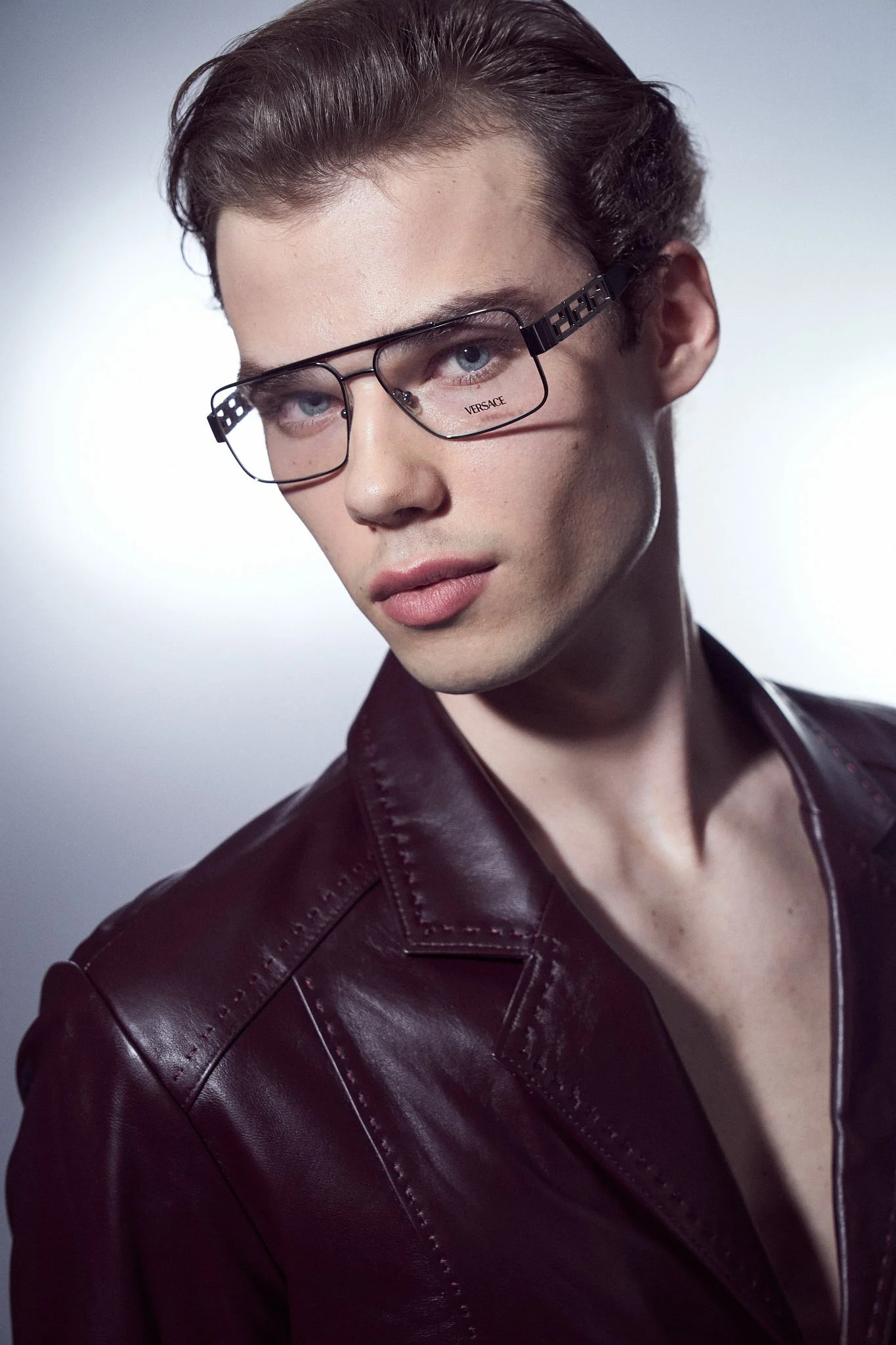 Studio campaign portrait of a male model wearing Versace designer eyeglasses and a dark leather jacket, lit in a sharp commercial fashion style.