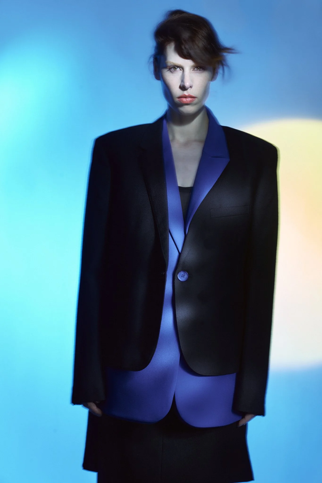 Full-body fashion editorial photograph of a model against a blue background, wearing a long black coat with a structured blue trim design.⁠