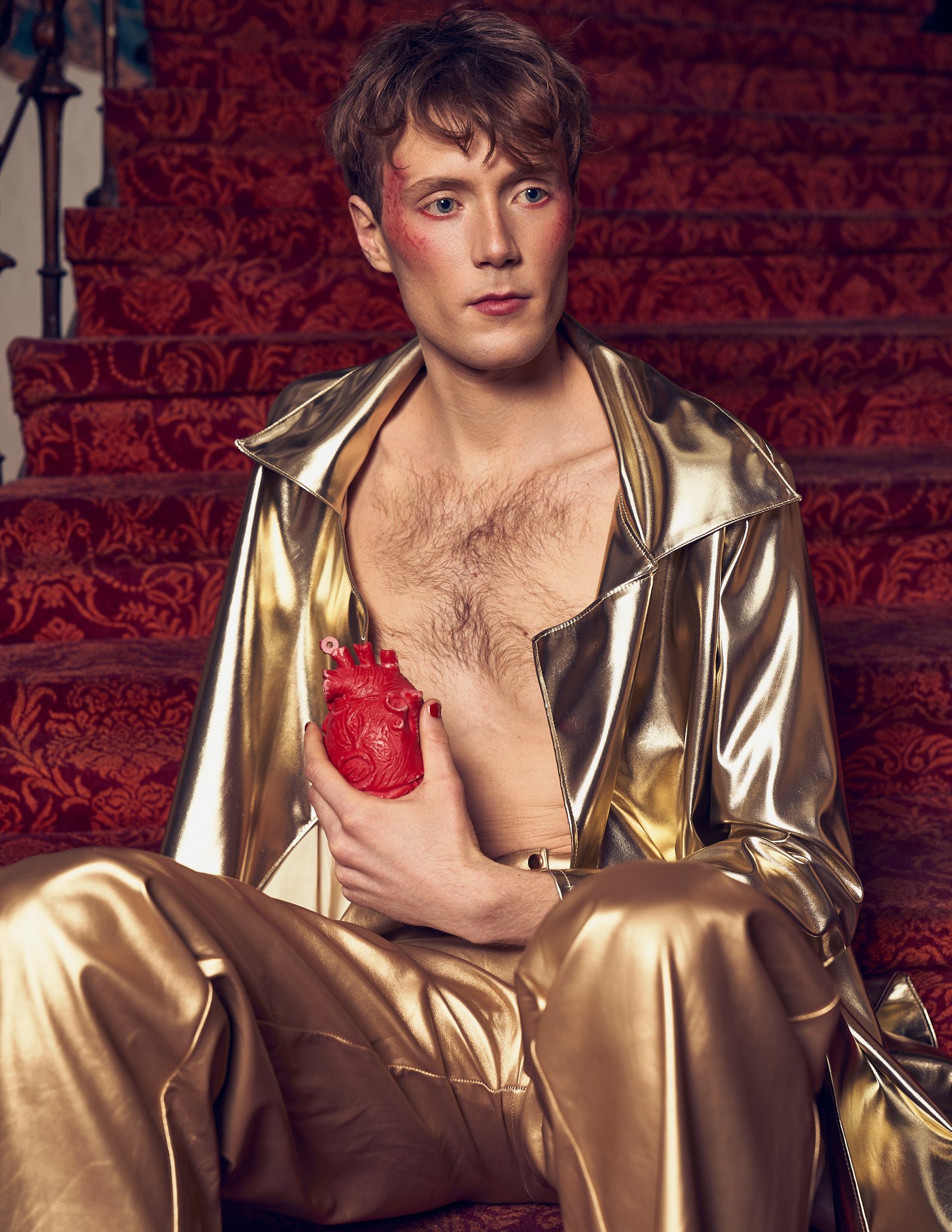 Location fashion portrait of a male model sitting open-chested on an ornate carpeted hotel staircase, wearing a metallic gold suit and holding a red anatomical heart sculpture.⁠