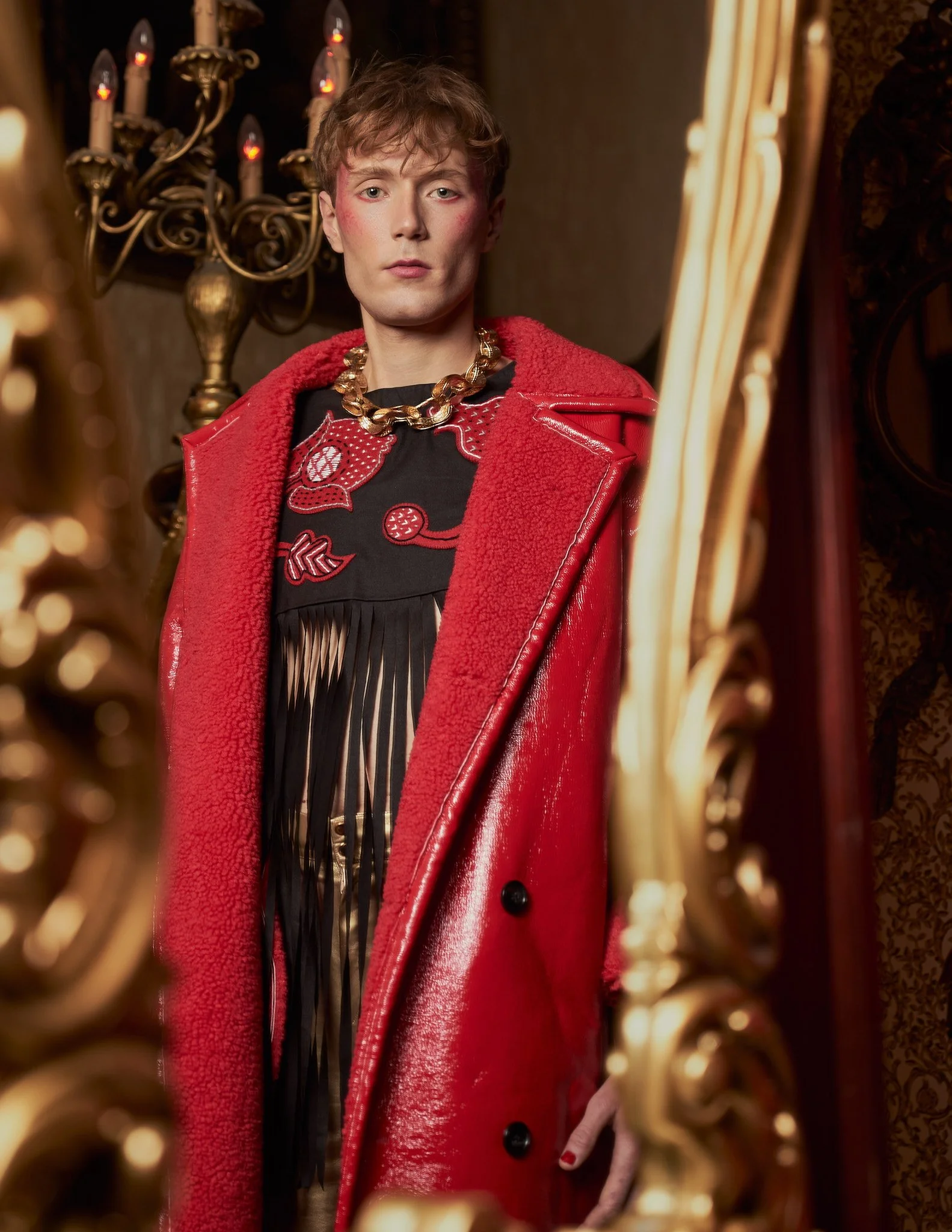 Editorial photograph captured through a gold framed mirror, showing a male model wearing a long textured red coat and ornate gold necklace.⁠