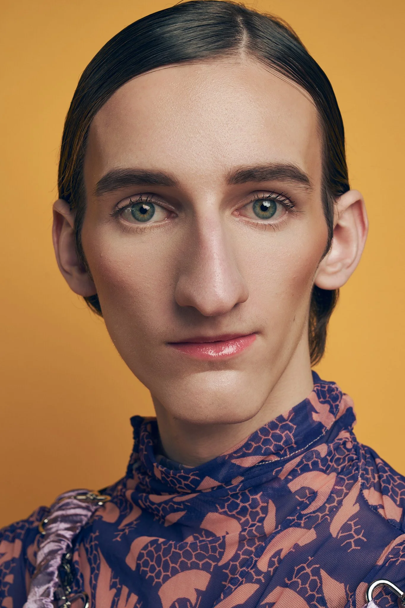 Close-up studio portrait of a male model with slicked hair, wearing a high-neck patterned blue and dark purple mesh top against a warm background.⁠