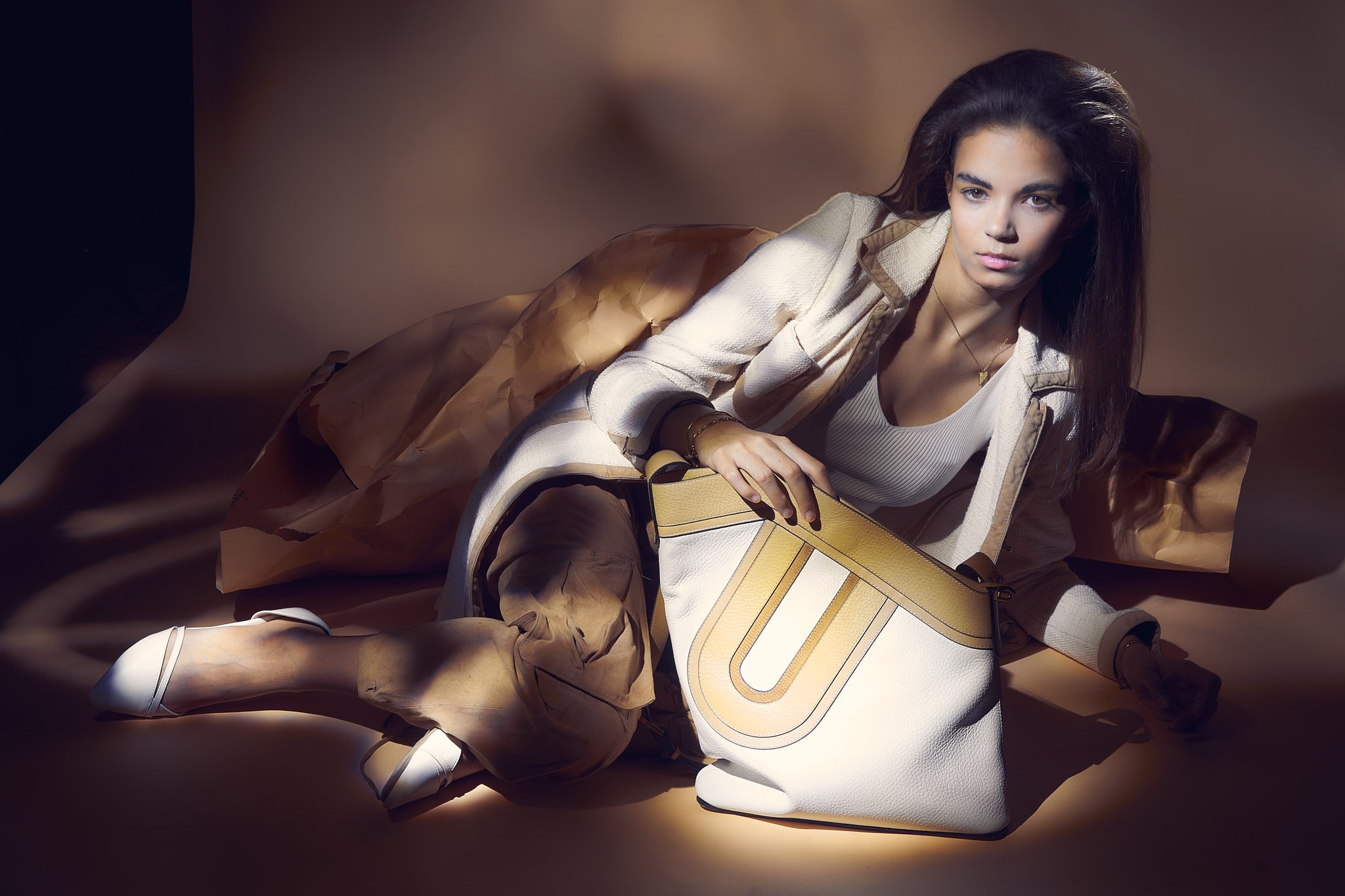 Fashion model reclining in studio light with a large white-and-camel designer handbag, photographed by Belgian fashion photographer in a warm beige setting.