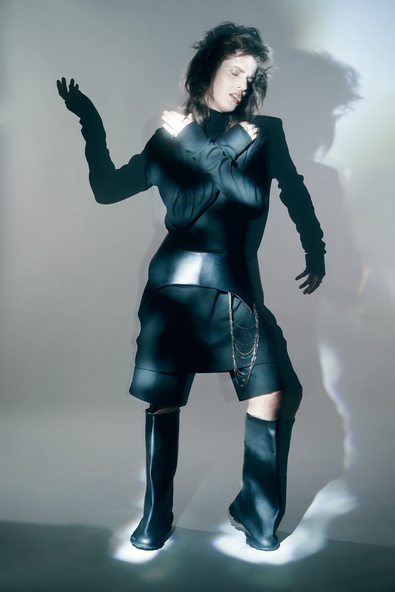 Full-body edgy menswear editorial shot of a model posing dynamically in a sharp black jacket, shorts, and tall leather boots under high-contrast studio lighting.⁠