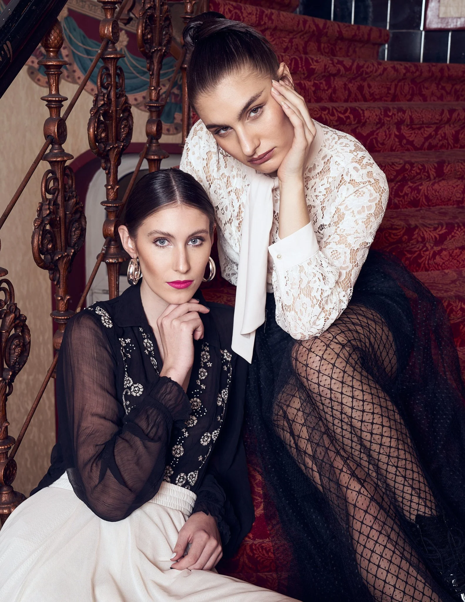 Fashion portrait of two female models posing on a carpeted staircase in lace blouses, sheer black tights, and vintage dark romantic styling.⁠
