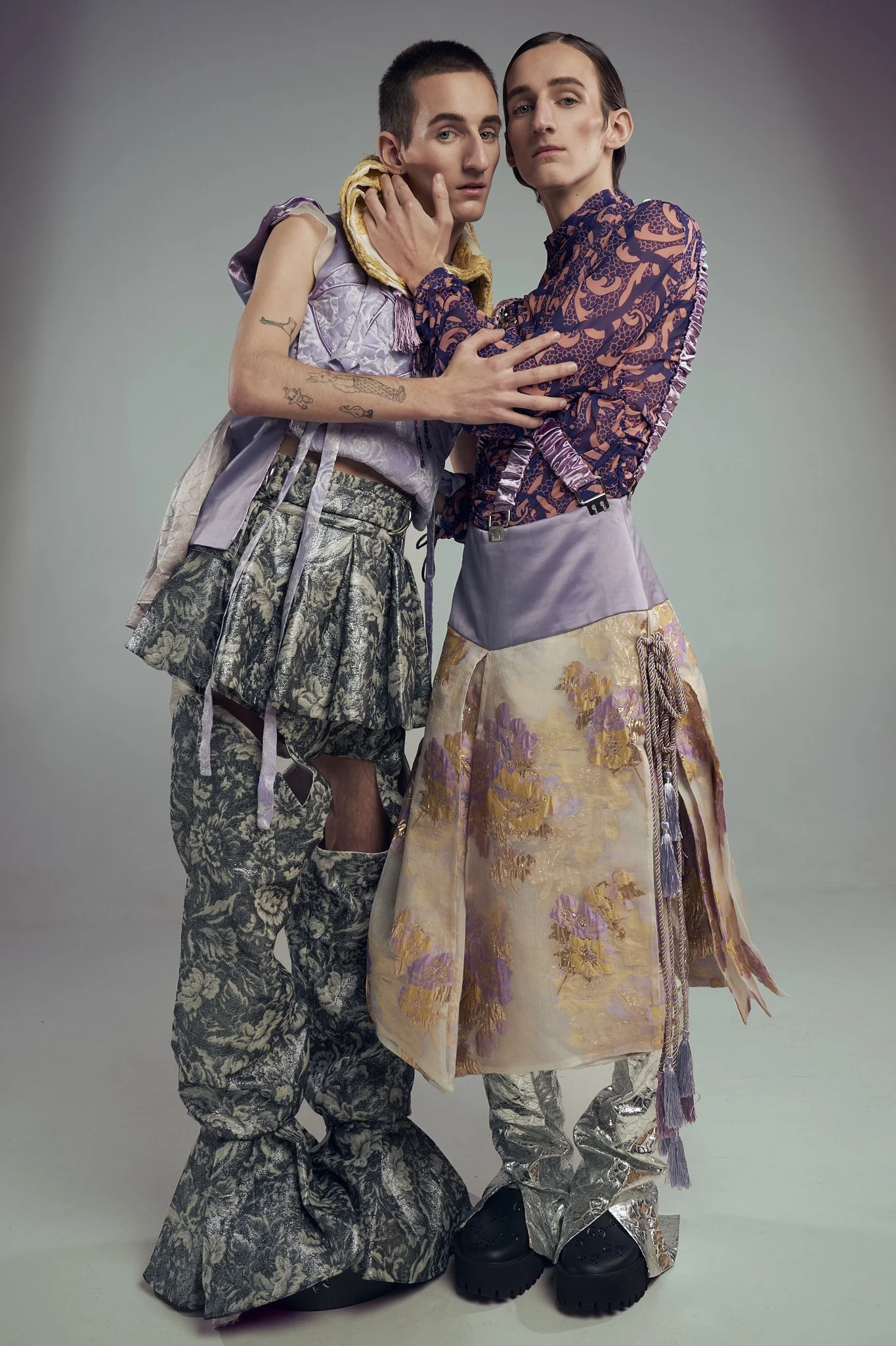 Full-body studio photograph of twin male models posing together in eclectic avant-garde tailoring, featuring patterned bell-bottom trousers and a brocade skirt overlay.⁠