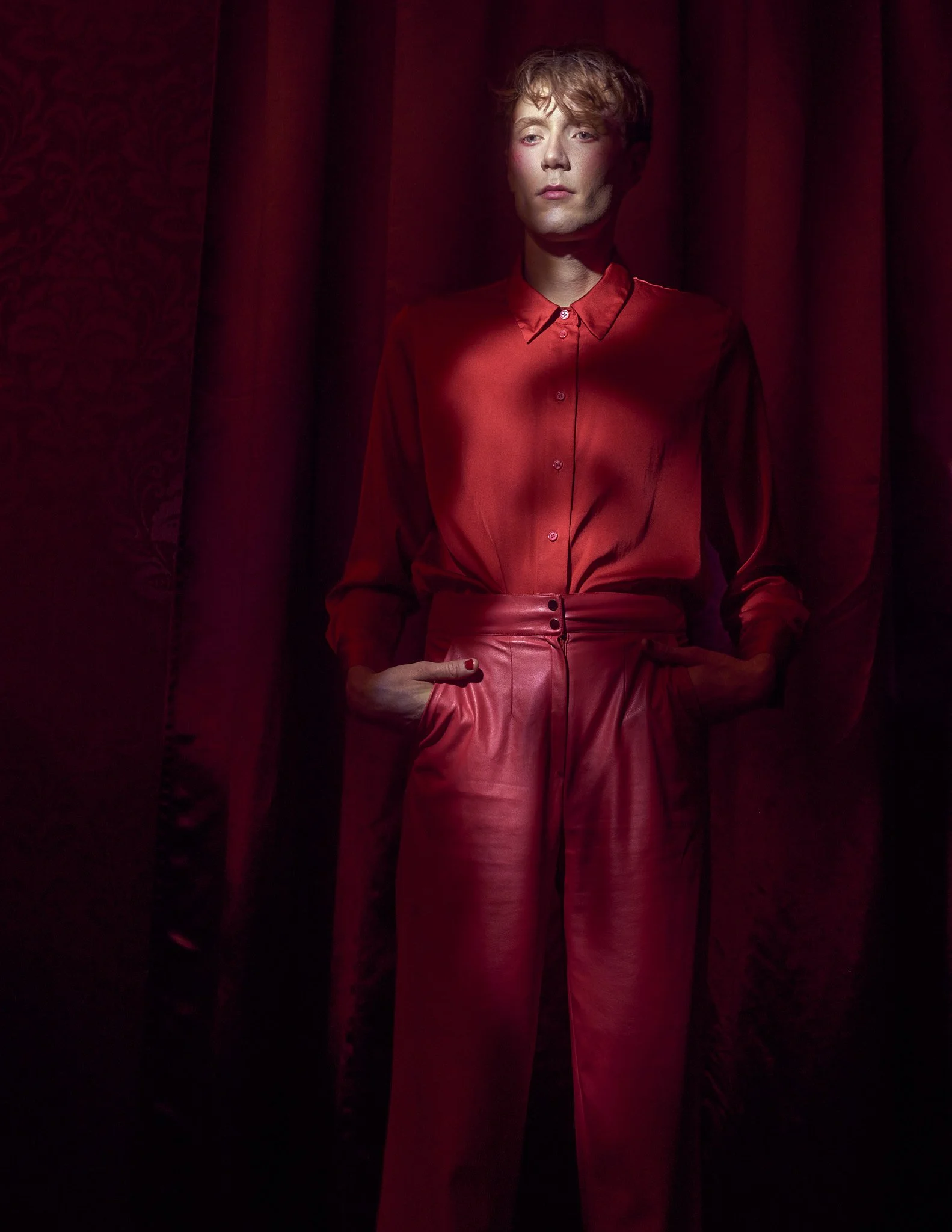 Full-length editorial photo of a male model dressed in a long-sleeved red silk top and wide-leg red trousers standing against a deep red velvet curtain.⁠