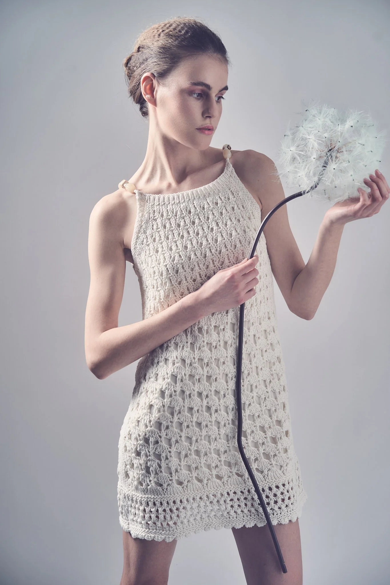 Full-length fashion editorial portrait of a model wearing a sleeveless white crochet minidress holding an oversized stem with a large dandelion head.⁠