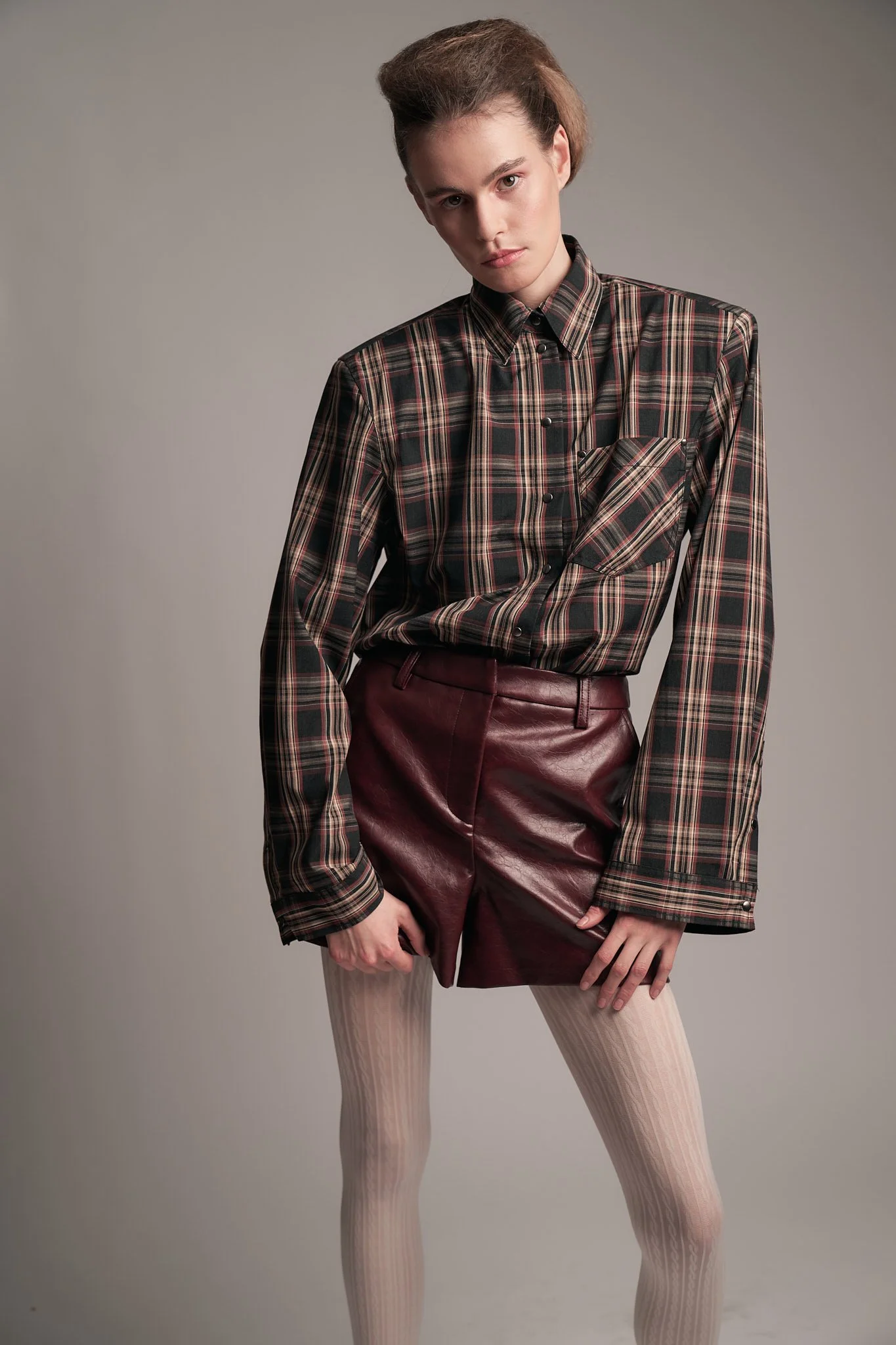 Three-quarter editorial shot of a model wearing a brown button-down plaid shirt tucked into deep burgundy leather trousers.⁠
