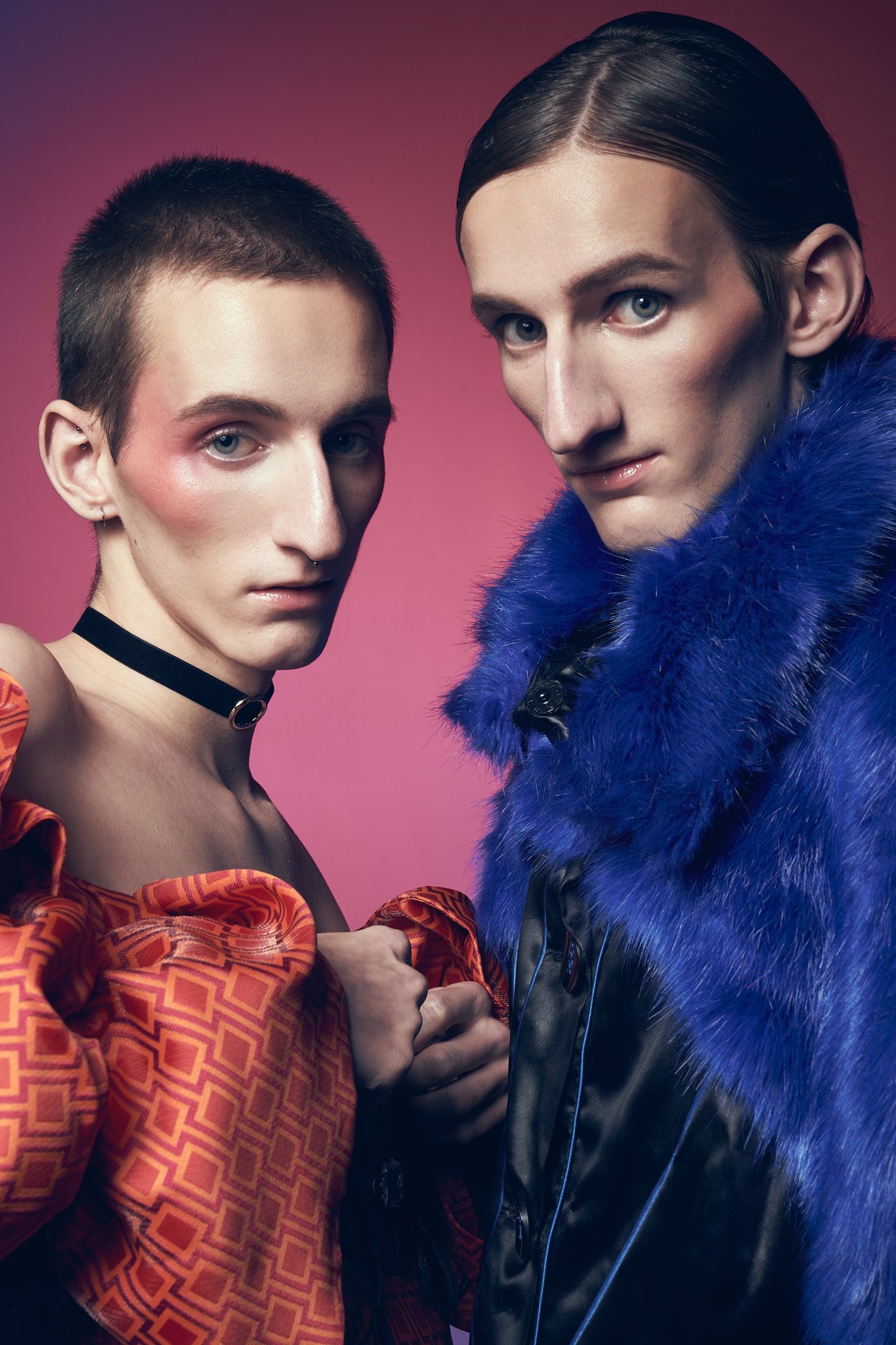 Editorial portrait of twin male models: one wearing an orange patterned off-the-shoulder outfit with a black choker, standing beside the other wearing a rich blue faux fur coat.⁠