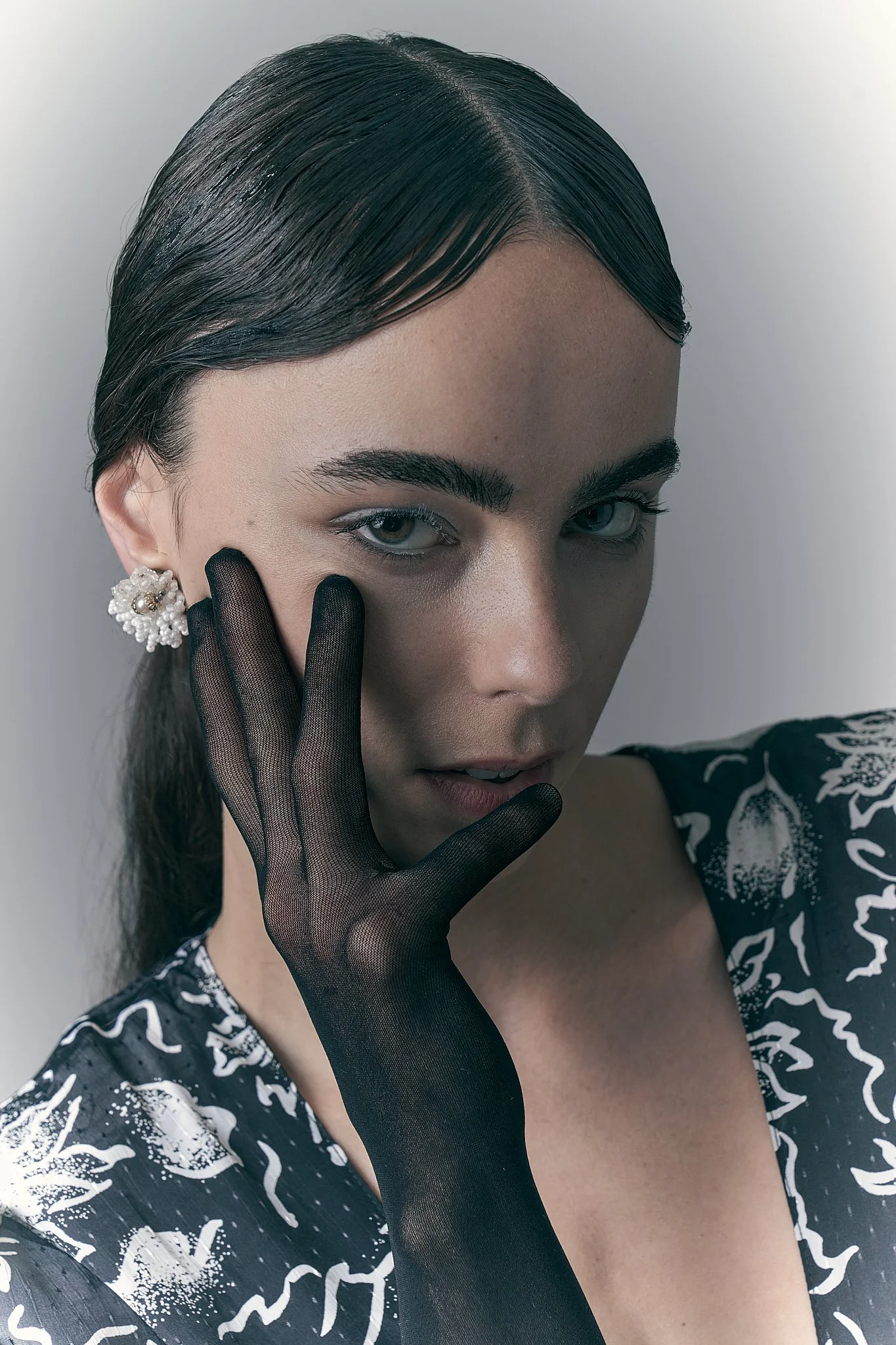 Close-up fashion portrait of a model with sleek hair wearing a black sheer glove over her face and a black-and-white floral print top.⁠