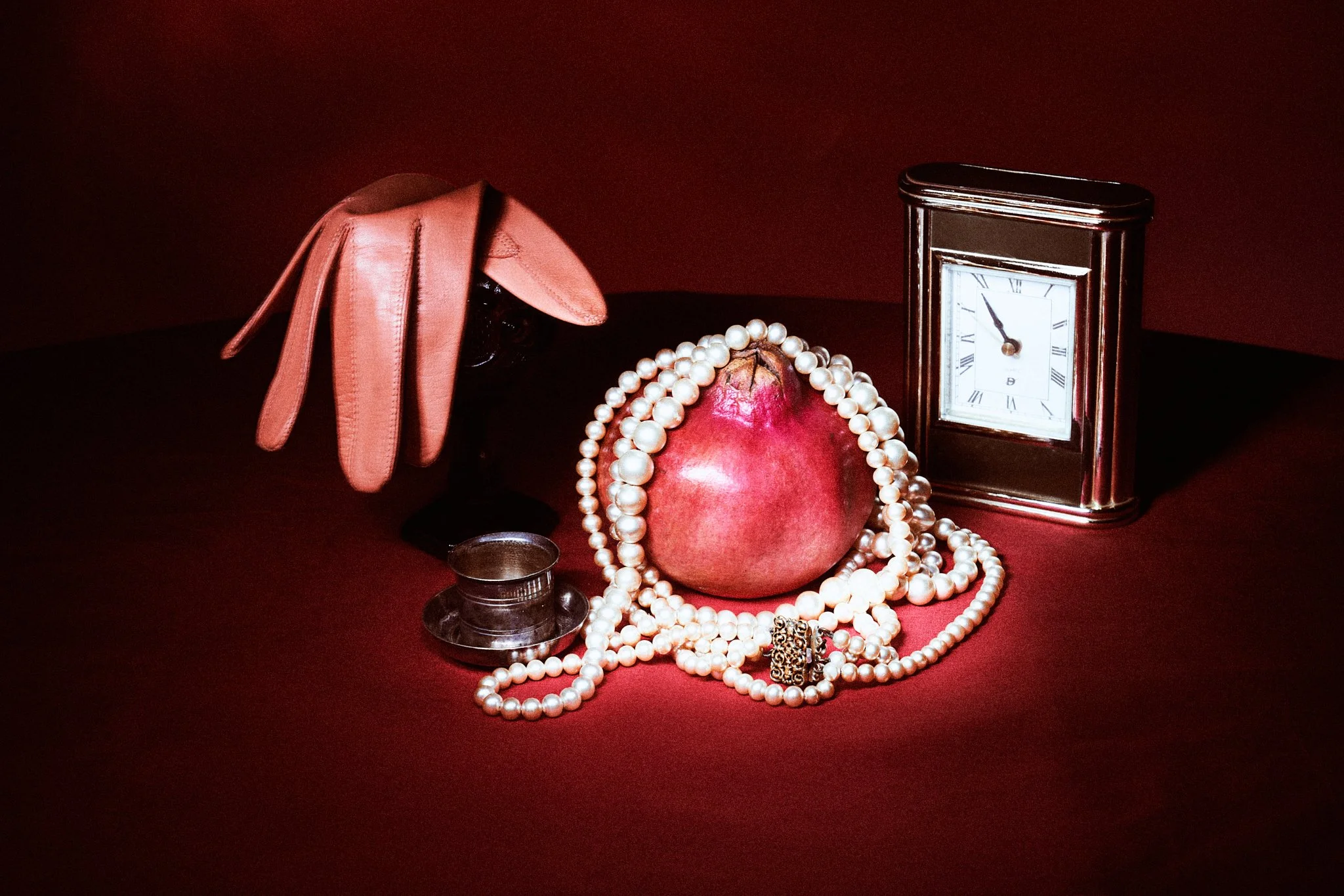 Still life studio photograph of a pomegranate wrapped in pearl necklaces alongside a small vintage table clock on a dark red surface.