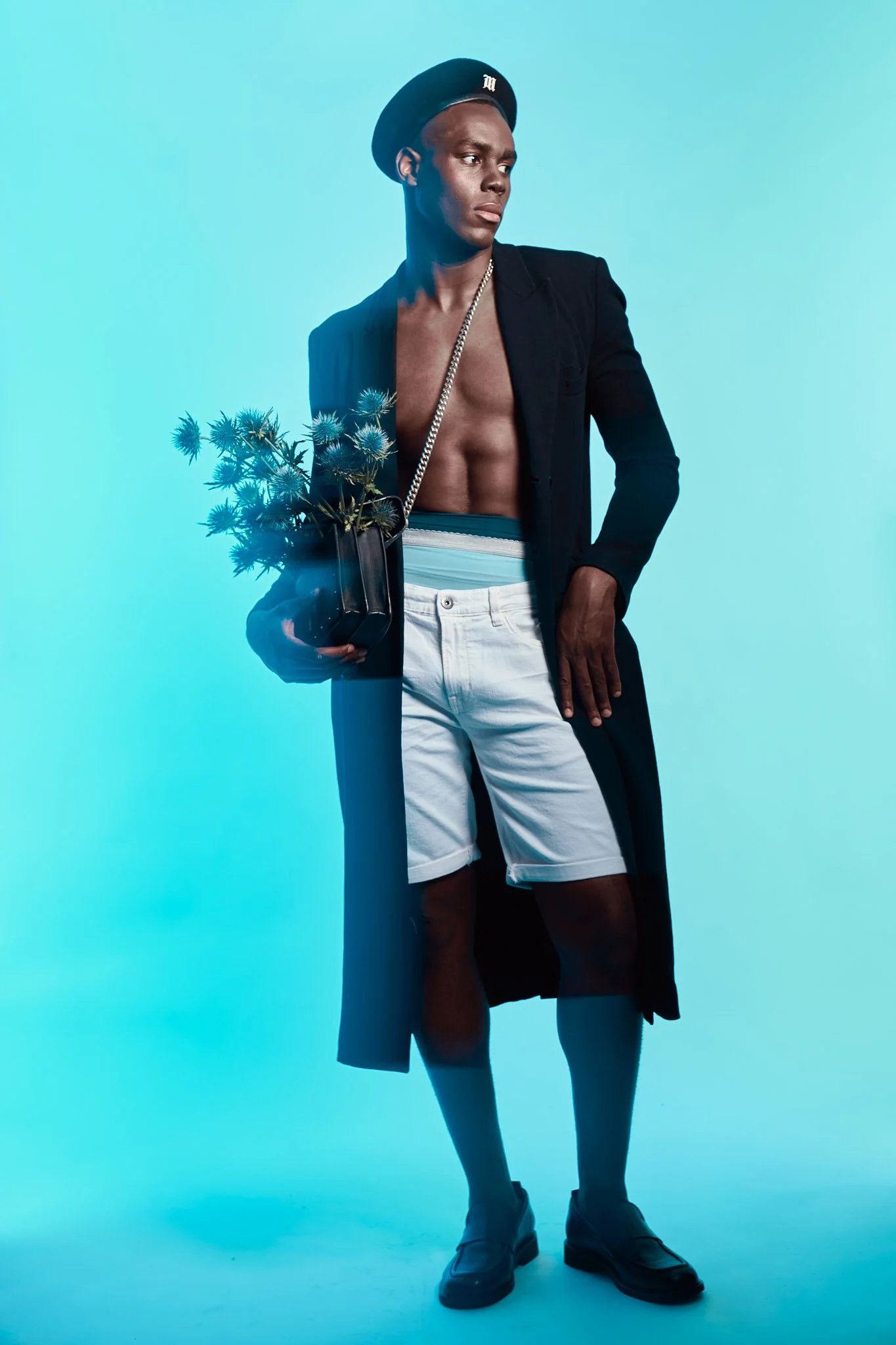 Full-body fashion photograph of a Black male model in a black beret, shirtless under a long black coat, white shorts, and blue socks against a solid blue backdrop, holding a bouquet of thistles.⁠