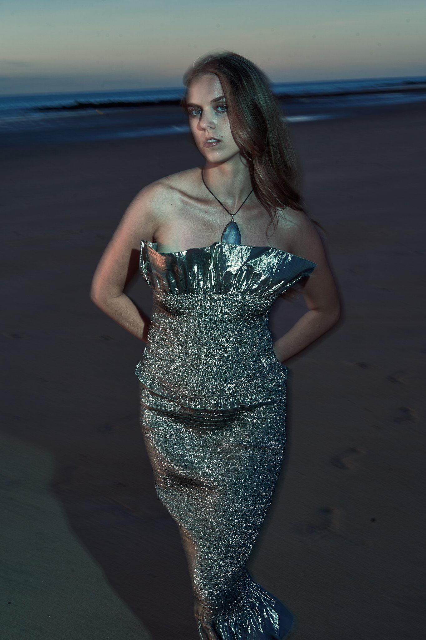 Woman in shimmering metallic strapless dress posing on beach at sunset, dramatic golden hour fashion photography by Belgian photographer Simon Rogghe