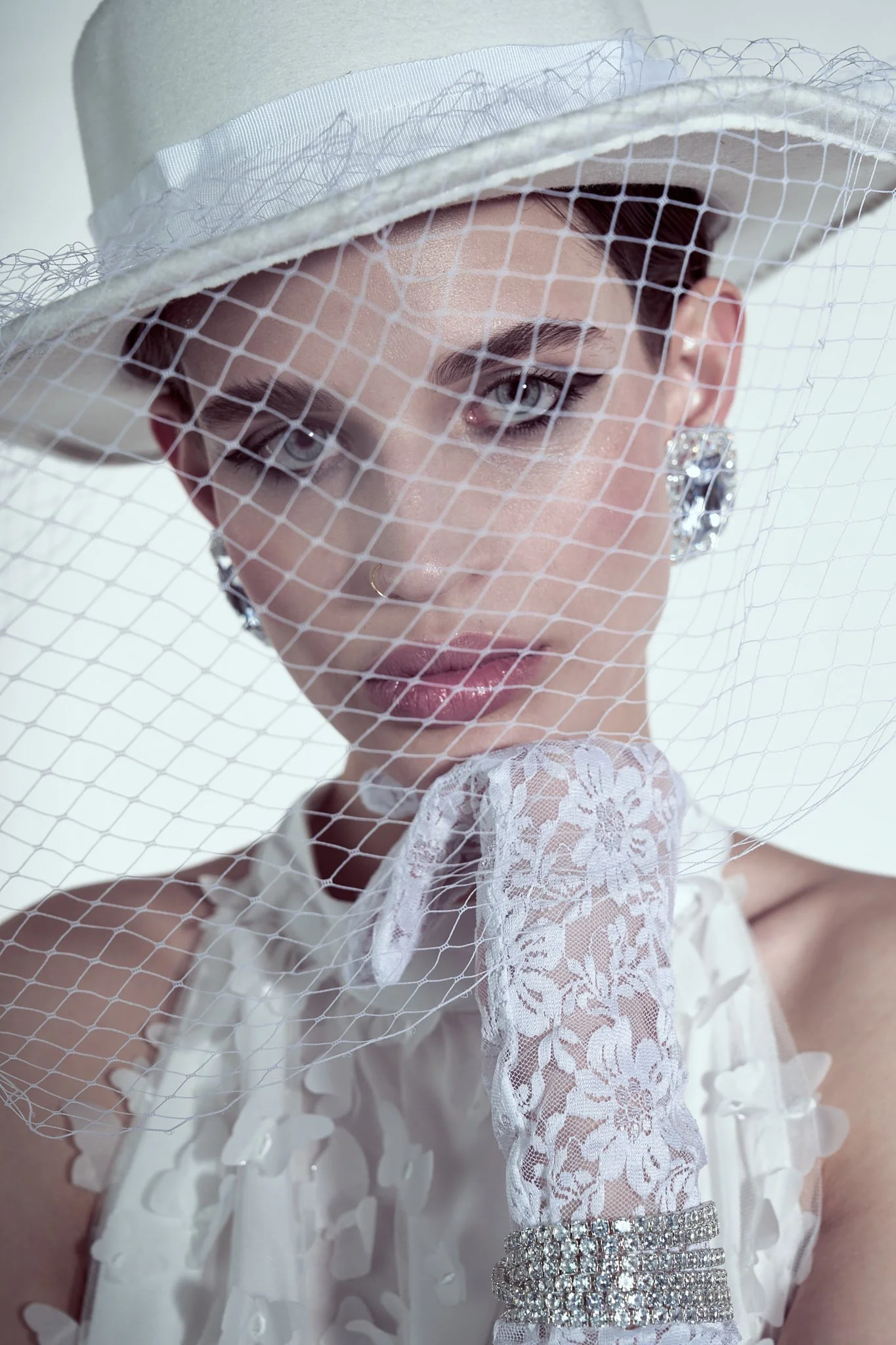 Close-up fashion portrait of a model in a white fedora hat with a netting birdcage veil, lace gloves, and crystal statement earrings.⁠