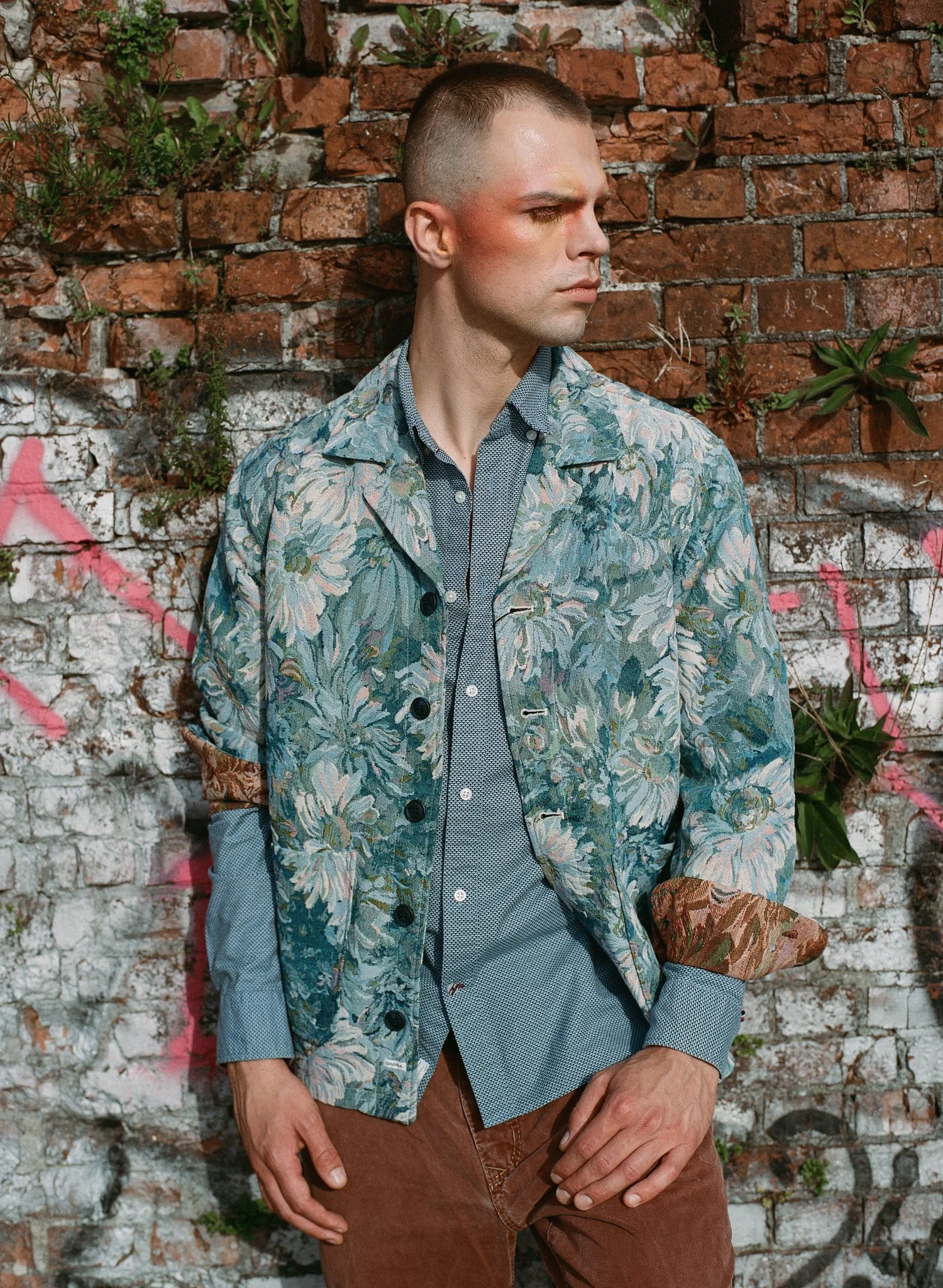 Fashion model wearing a floral-patterned jacket and blue shirt standing against a weathered brick wall with dramatic natural light and editorial styling. Shot on Portra 400 on analog medium format with a Mamiya 645.