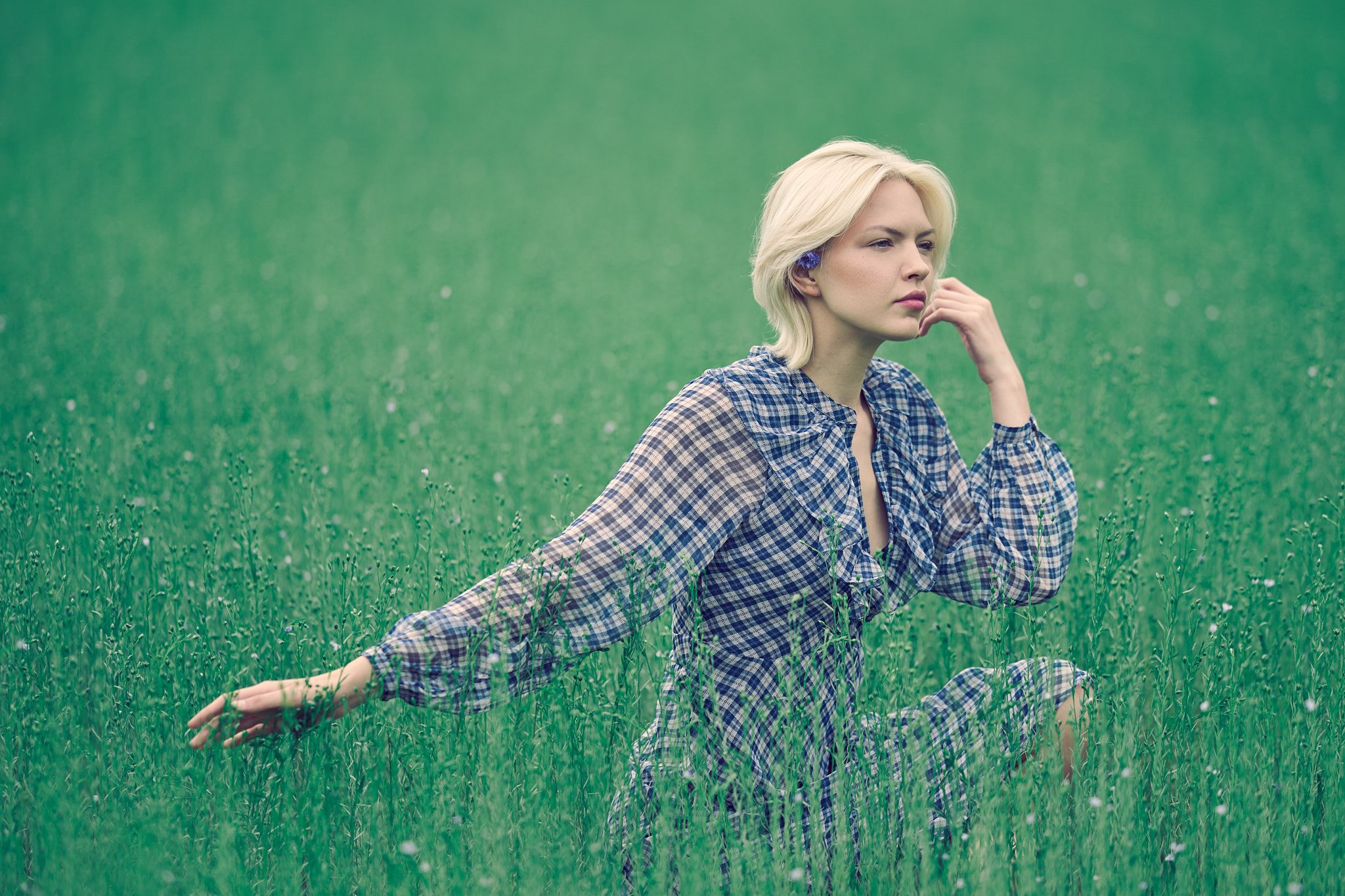  A blonde woman wearing a blue and white plaid dress sits in a lush green field resting her chin on her hand.