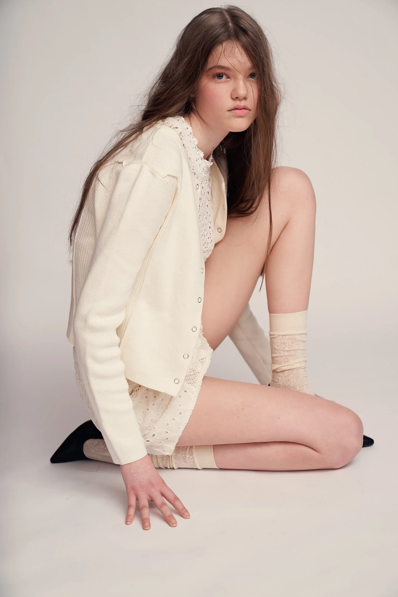 Soft campaign portrait of a young model kneeling in a monochrome cream outfit with lace textures, styled in minimalist neutrals—studio fashion photography by a Belgian photographer based in Ghent.