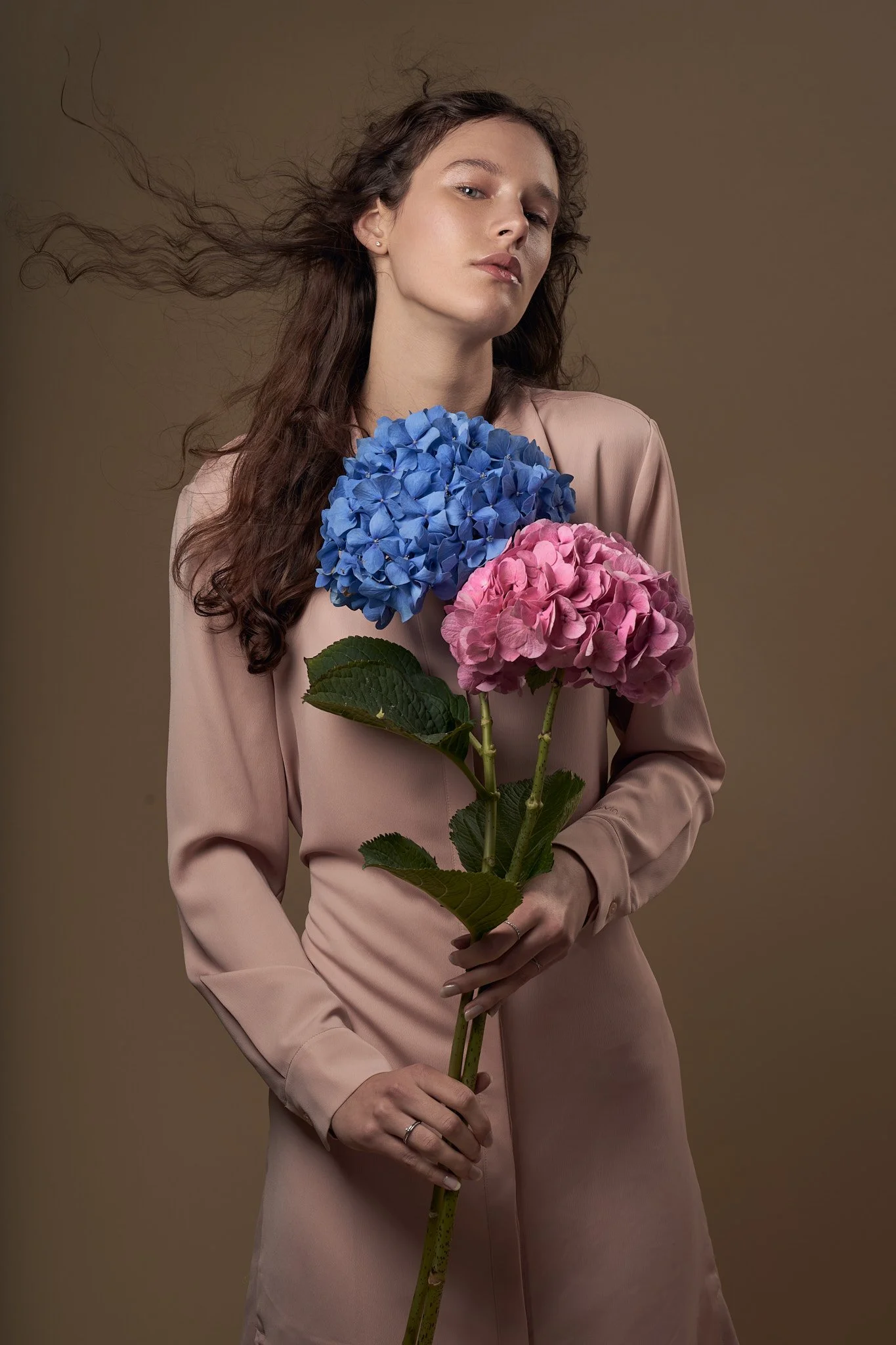 Editorial portrait of a woman in a neutral long-sleeve dress holding bright blue and pink hydrangea flowers against a warm studio backdrop.