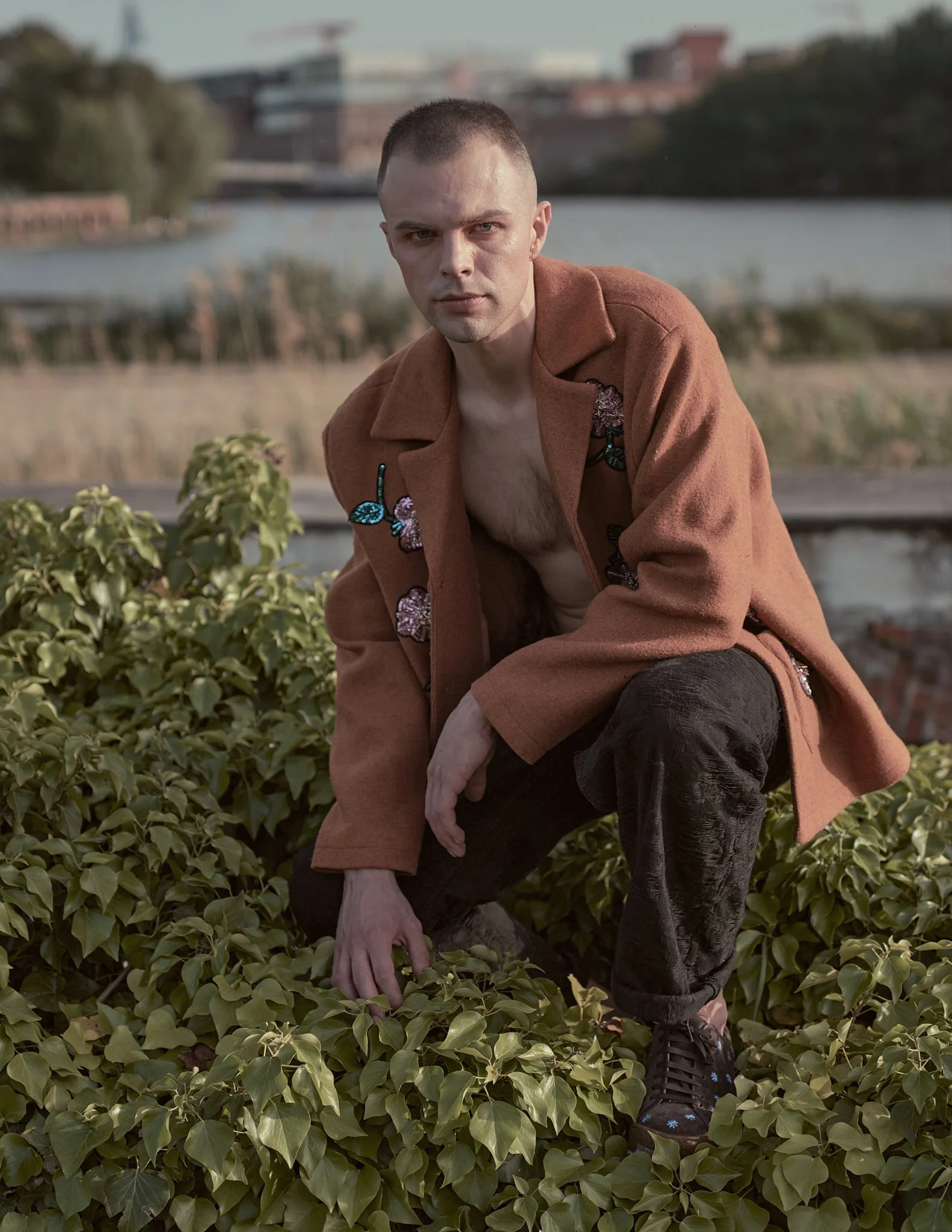 Editorial fashion portrait of a young man in an open terracotta wool coat with floral brooches, crouching among green foliage at an industrial waterfront location in Ghent, Belgium.