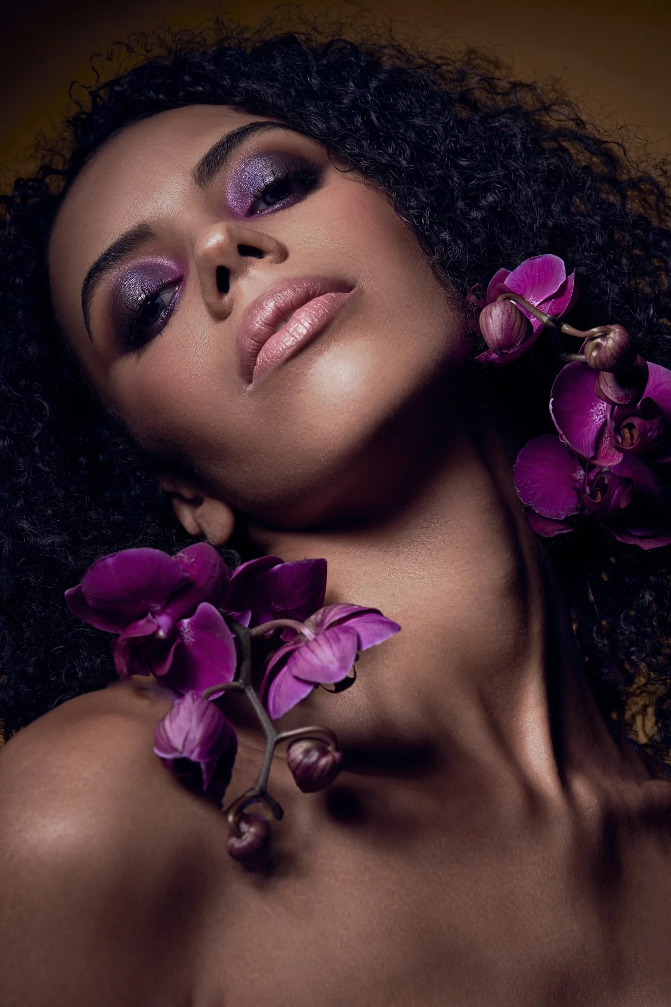 Beauty portrait of a woman with dark curly hair and vibrant purple eyeshadow, styled with deep purple orchid flowers.