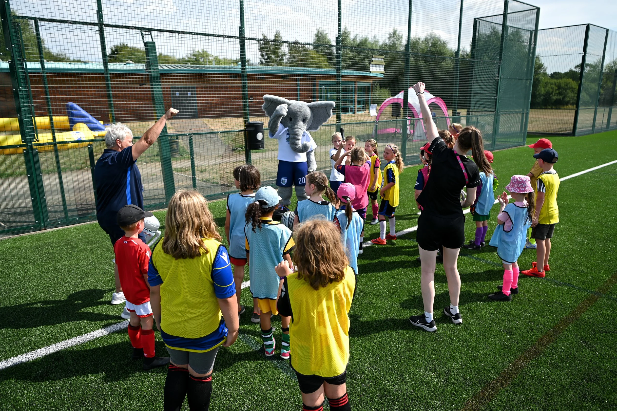 Children on a soccer field listening to a woman and a person in an elephant mascot costume, with some children raising their hands, near a fenced playground. Shot for Nottinghamshire Football Association by Shaun Hardwick (trading as S.H Photos).