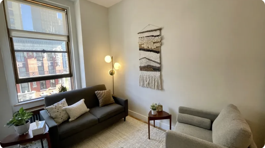 Therapy office with a window, a black sofa with decorative pillows, a gray armchair, a small side table with plants, a floor lamp, and wall art. In-person Accelerated Resolution Therapy in New York City with Laura Geftman, LCSW.