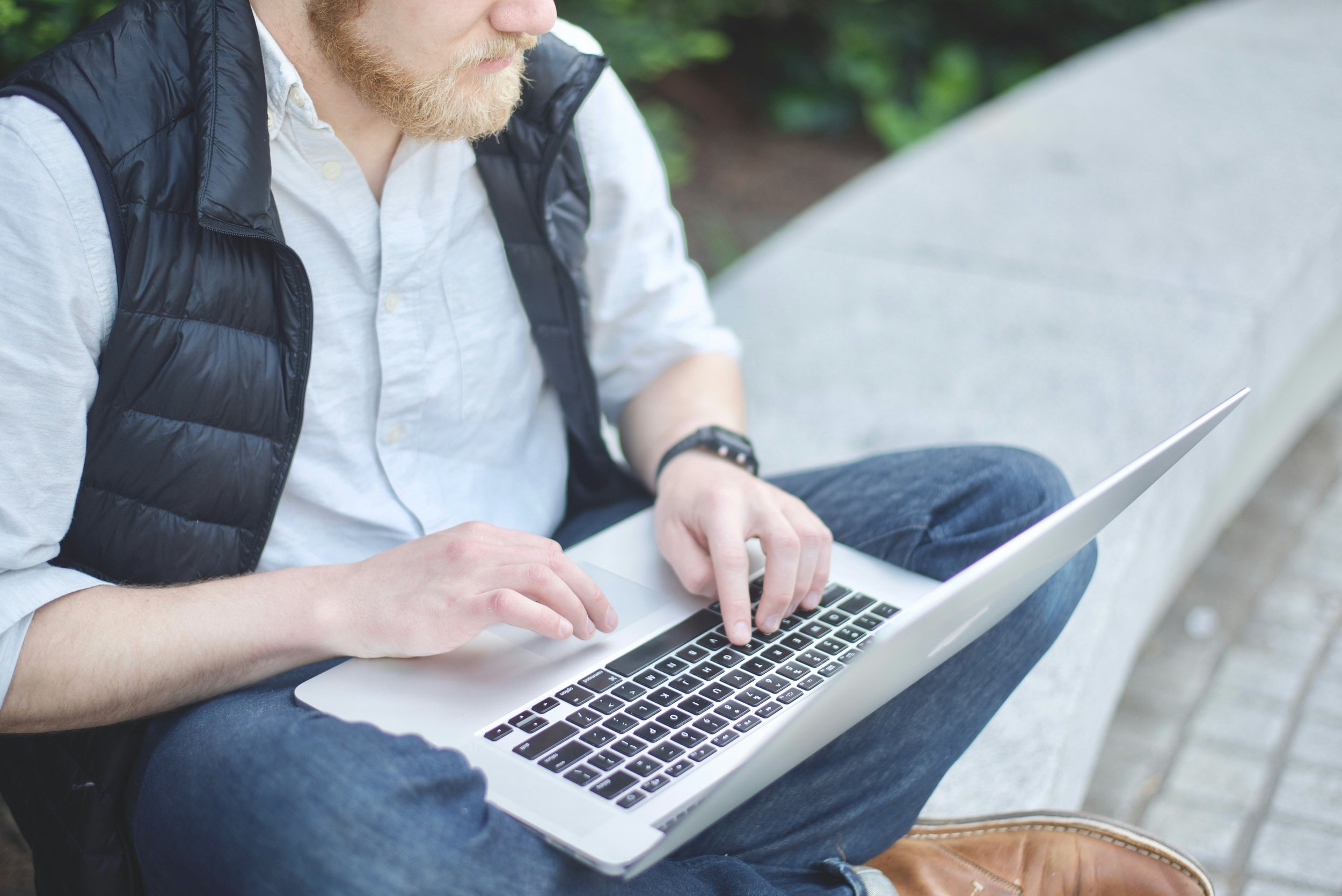 A man with a beard sitting on a park bench using a laptop, dressed in a white shirt, black vest, and jeans. Accelerated healing for adults who want fast results—not just talk. In-person therapy in Philadelphia & online throughout PA, NY, NJ, & FL.