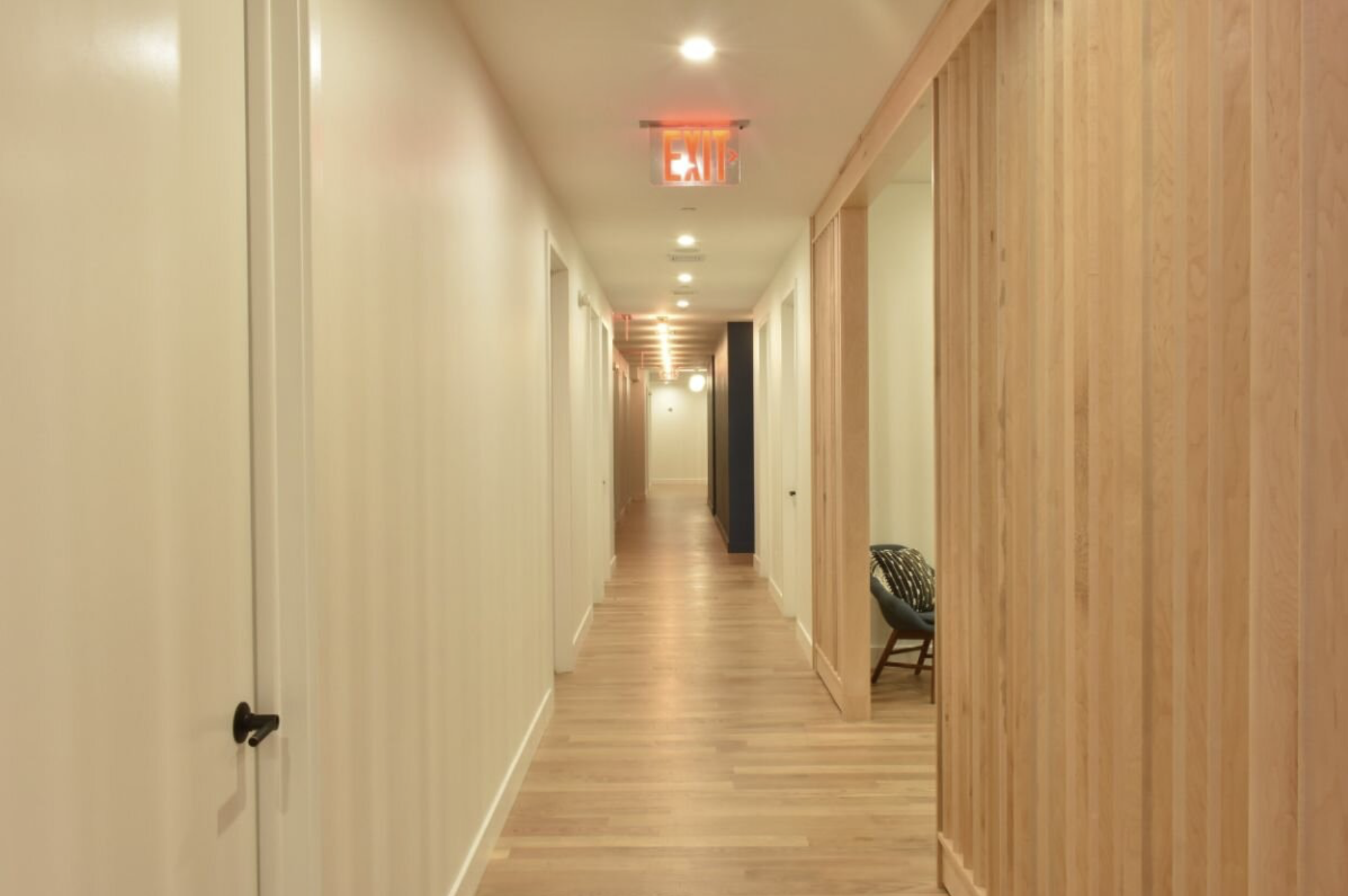 The hallway with wooden flooring, cream-colored walls, several doors, and a lit red exit sign overhead. In-person Accelerated Resolution Therapy in New York City with Laura Geftman, LCSW.