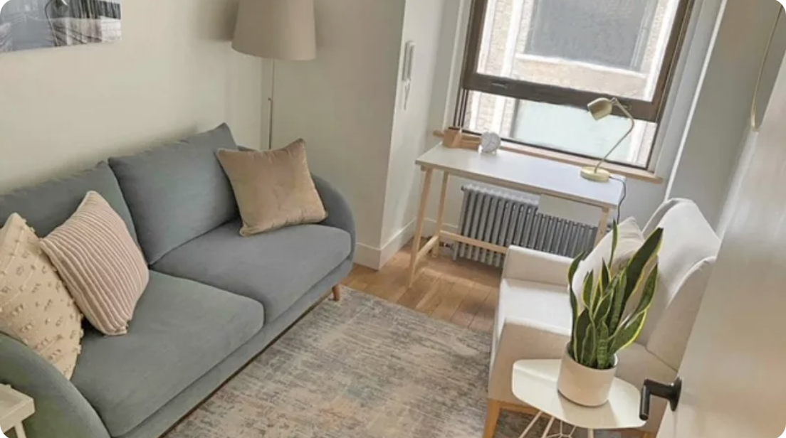 Cozy therapy office with gray sofa, beige pillows, white armchair, small white table with plant, wooden desk beside window, and hardwood floor. In-person Accelerated Resolution Therapy in New York City with Laura Geftman, LCSW.