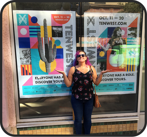 A woman standing in front of a colorful event poster at TENWEST Festival, wearing sunglasses, a floral top, and jeans, smiling and gesturing with her hands.