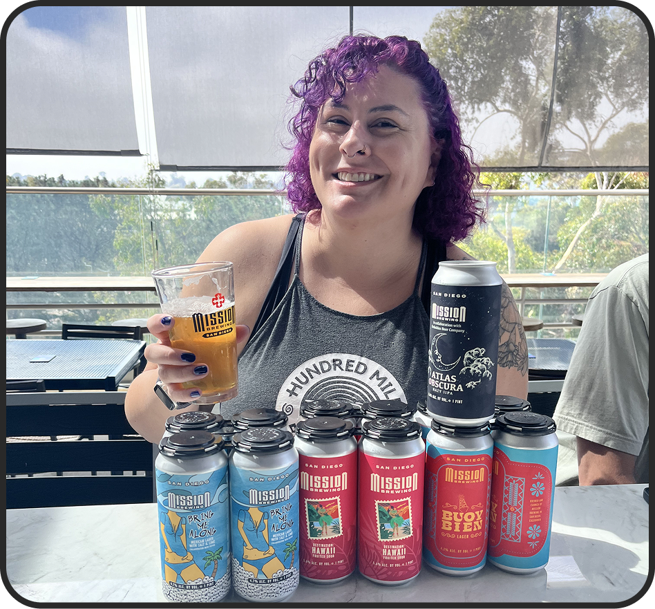 A woman with purple curly hair smiling and holding a glass of beer at a table with multiple colorful cans of Mission Brewing beers in front of her.