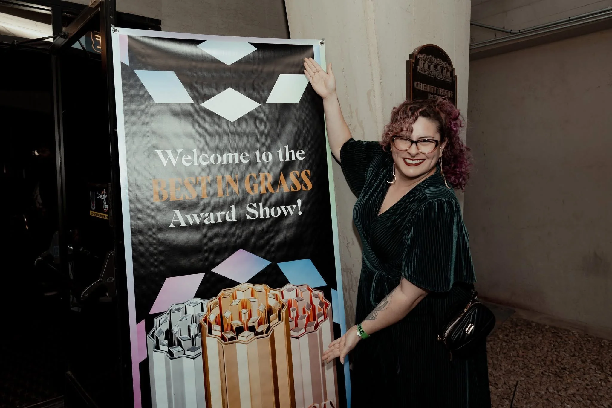 Woman with curly hair, glasses, and earrings smiling and pointing at a sign that reads 'Welcome to the Best in Grass Award Show.' She is standing indoors next to a decorative wall and a poster featuring a honeycomb pattern.