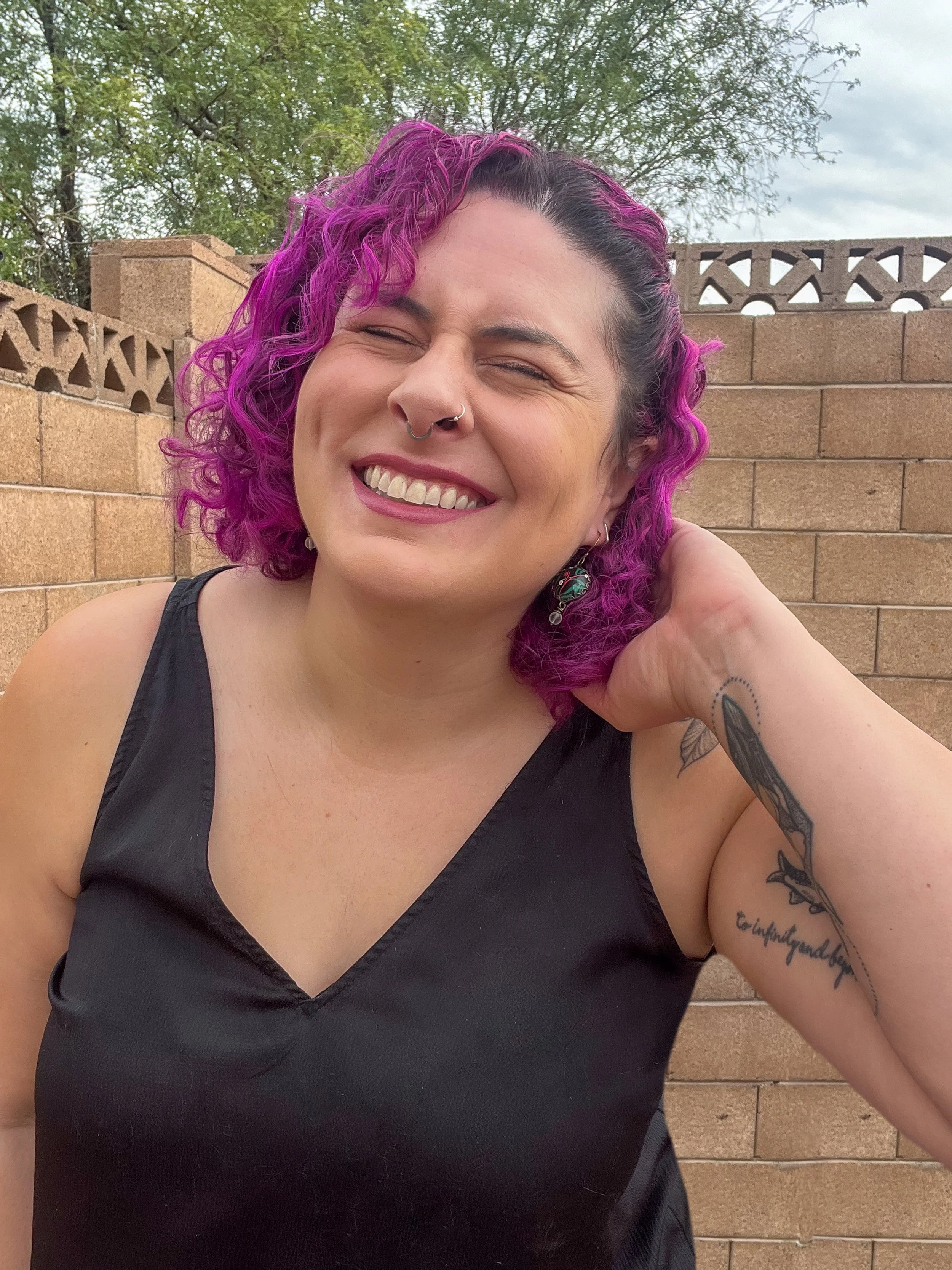 A woman with curly purple hair, wearing a black top, smiling with her eyes closed, outside with a brick wall and trees in the background.