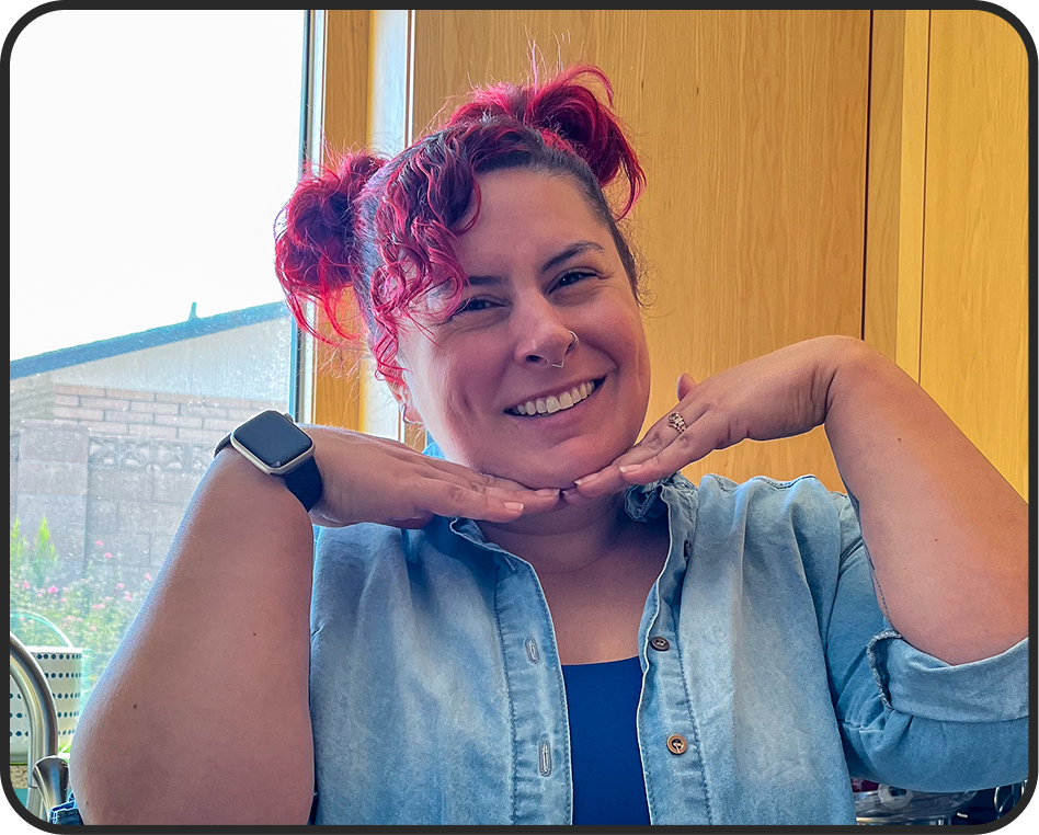 A smiling woman with pink curly hair styled in two buns, wearing a denim shirt, a black top, a smartwatch, and a ring, posing with her hands under her chin inside a room with wooden paneling and a window showing outside scenery.