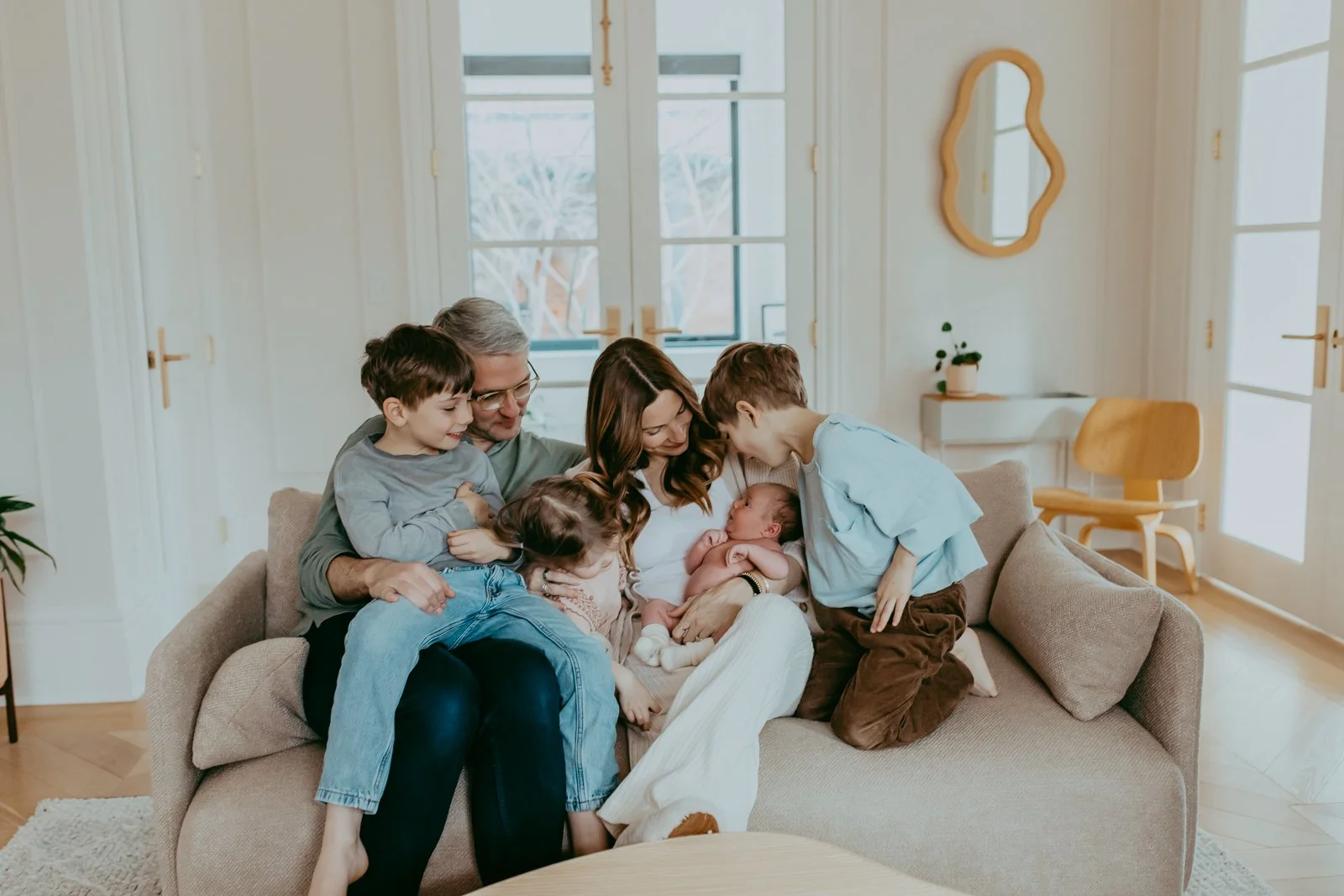 A family of seven sitting on a sofa, holding and admiring a newborn baby, in a well-lit living room with large windows, wooden floors, and modern decor.