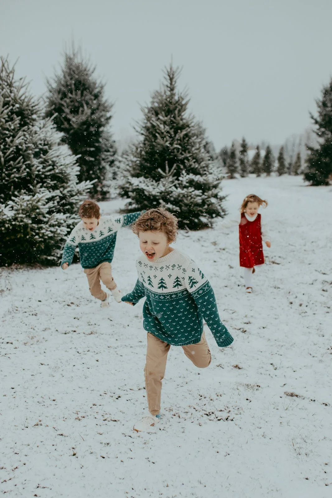 Three children playing and running in the snow near Christmas trees, wearing festive sweaters and a red dress outdoors.