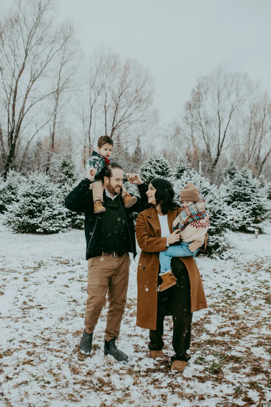 A family of four enjoying a snowy day outdoors. The father carries a young boy on his shoulders, while the mother holds a girl. They are standing on snow-covered ground with evergreen trees in the background.